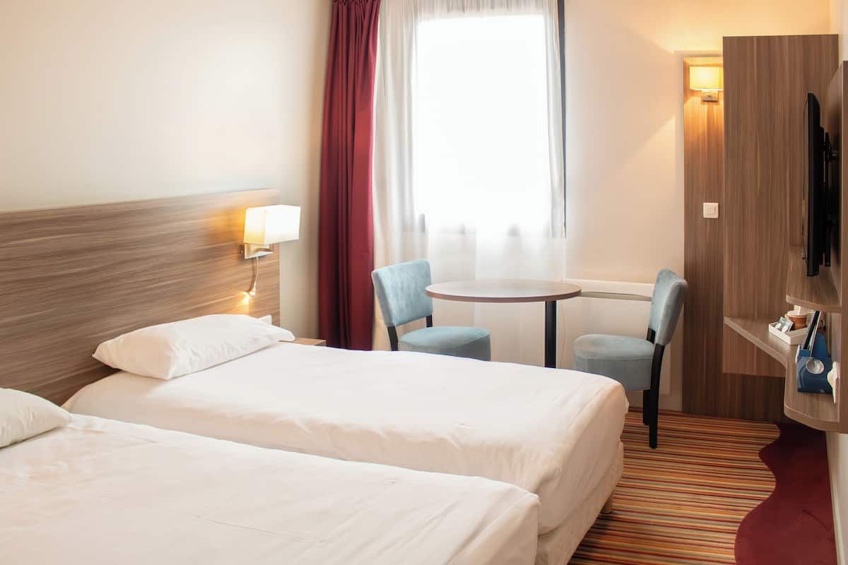 A hotel room with two beds, a small table, two chairs, a TV, and a window with curtains.