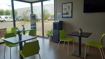 A hotel lobby with green chairs and tables, a large window offering a view of the street, and a poster advertising the hotel.