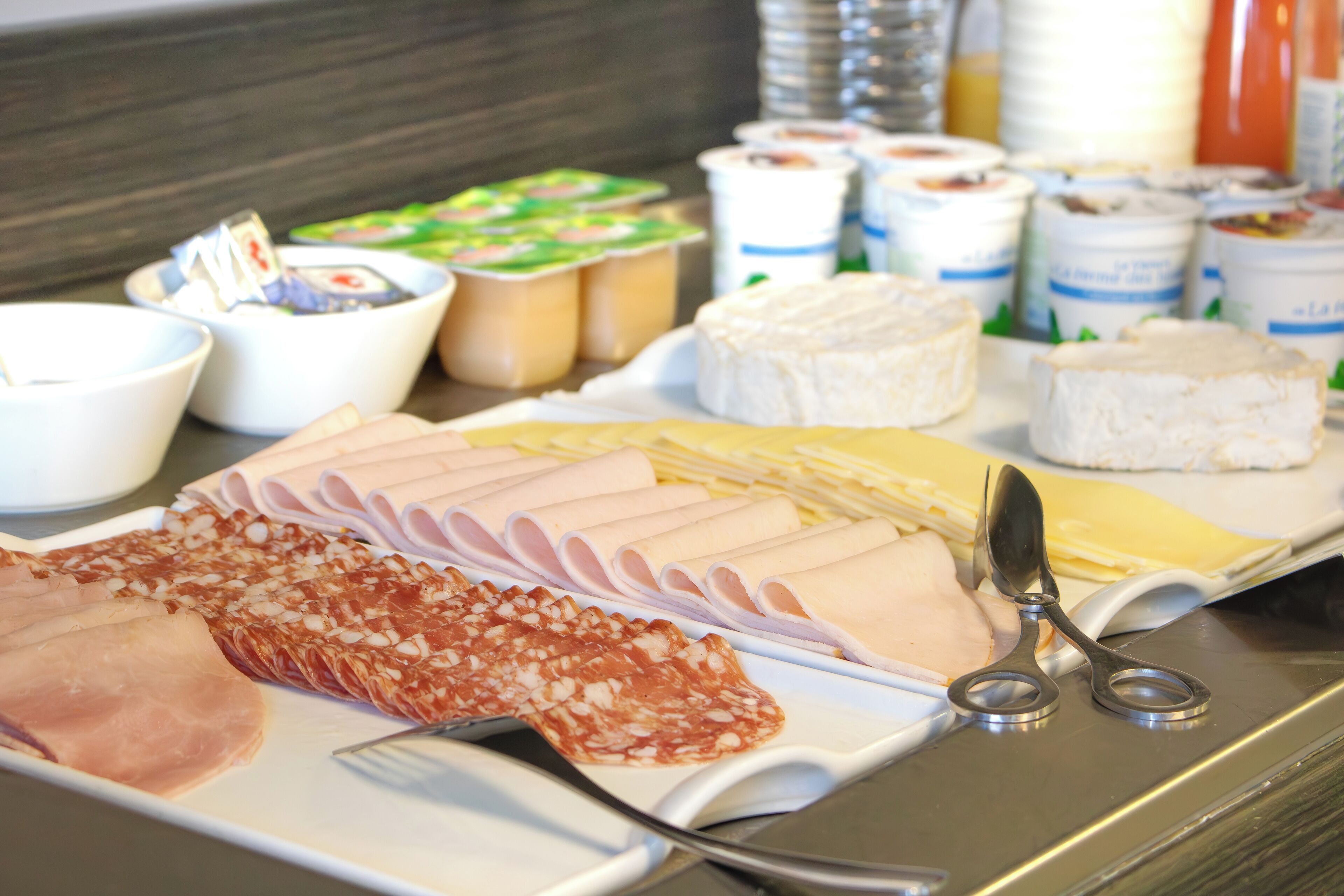 A buffet with various cheeses, meats, and yogurt containers.