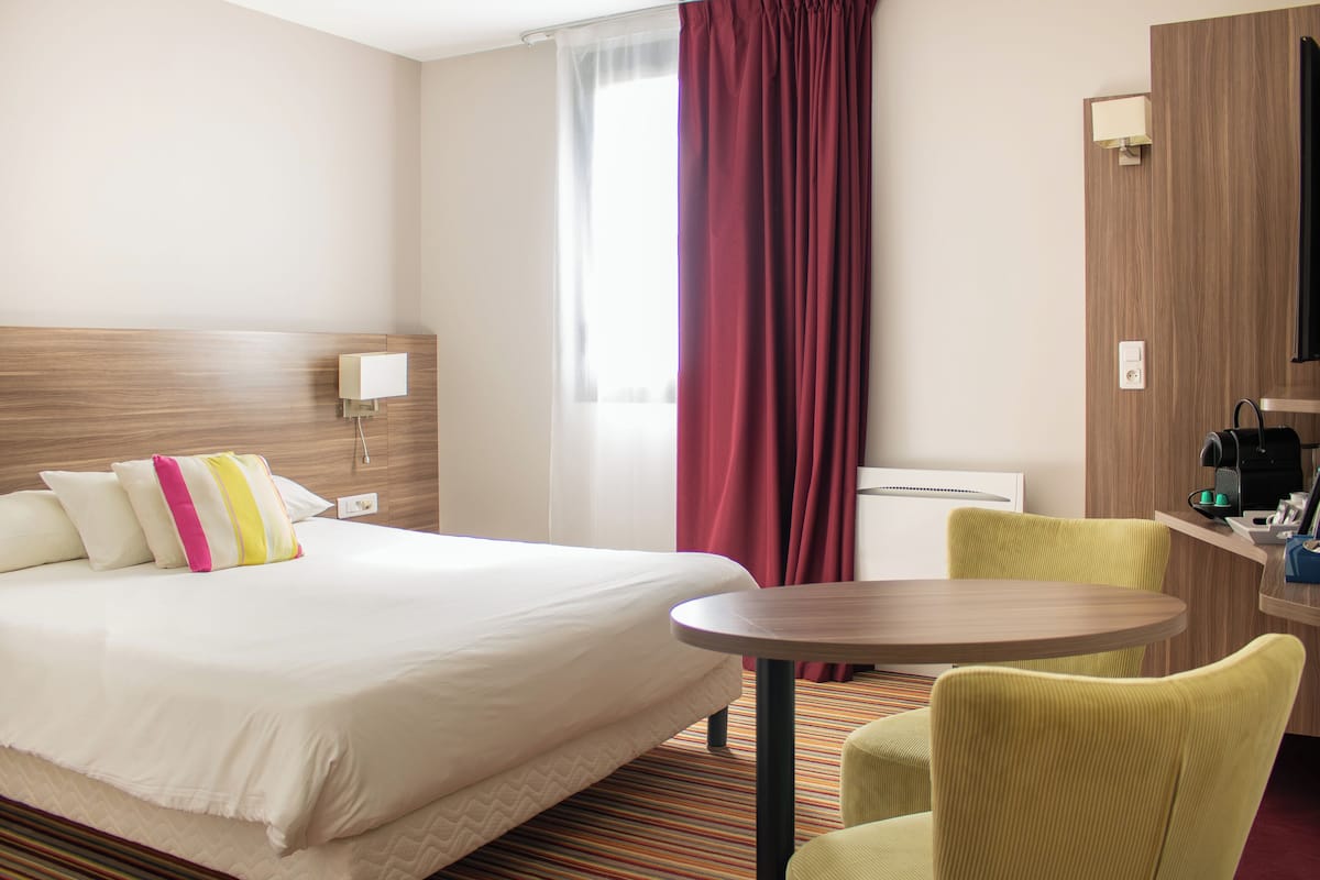 A hotel room with a bed, a round table, a chair, a window with curtains, and a wall-mounted lamp.