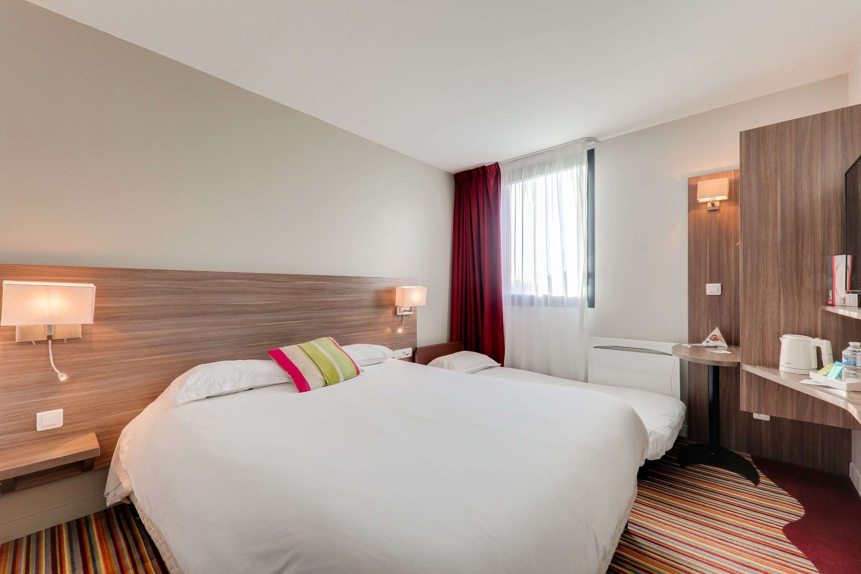 Habitación de hotel con una cama grande, dos lámparas de noche, un cabecero de madera, un escritorio pequeño con una lámpara, una cafetera y un tapete a rayas.