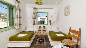 A small, cozy room with two single beds, a wooden chair, a small table with a cup, and a window with curtains. - Elounda Apartments (Agios Nikolaos)