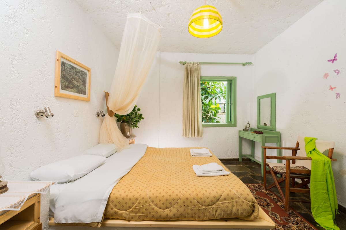 A bedroom with a bed, a desk, a chair, a hanging plant, and a window with a view of greenery.