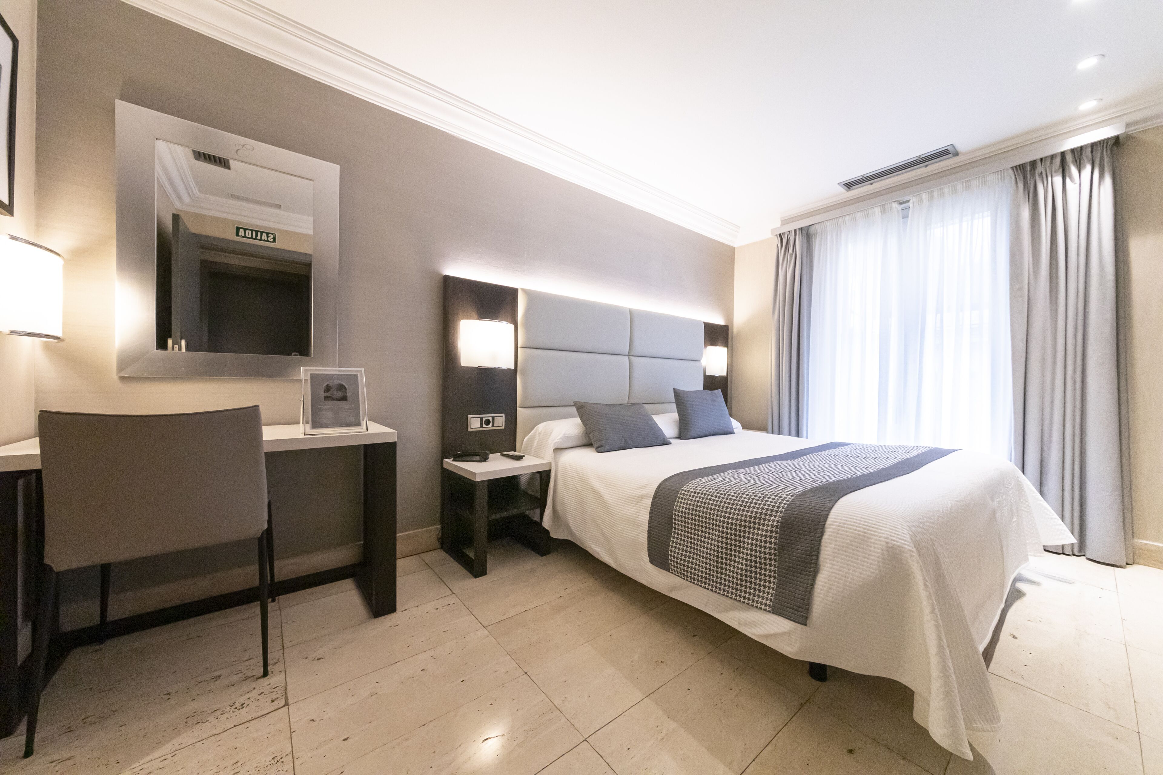 Habitación de hotel con cama, escritorio, silla, mesita de noche, espejo y ventana con cortinas.
