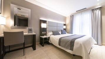 Habitación de hotel con cama, escritorio, silla, mesita de noche, espejo y ventana con cortinas.