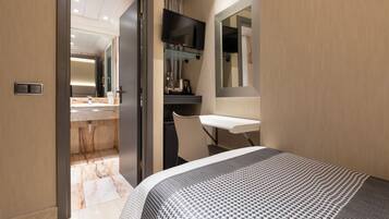 A modern hotel room with a bed, a desk, a chair, a television, and a bathroom with a mirror and sink.