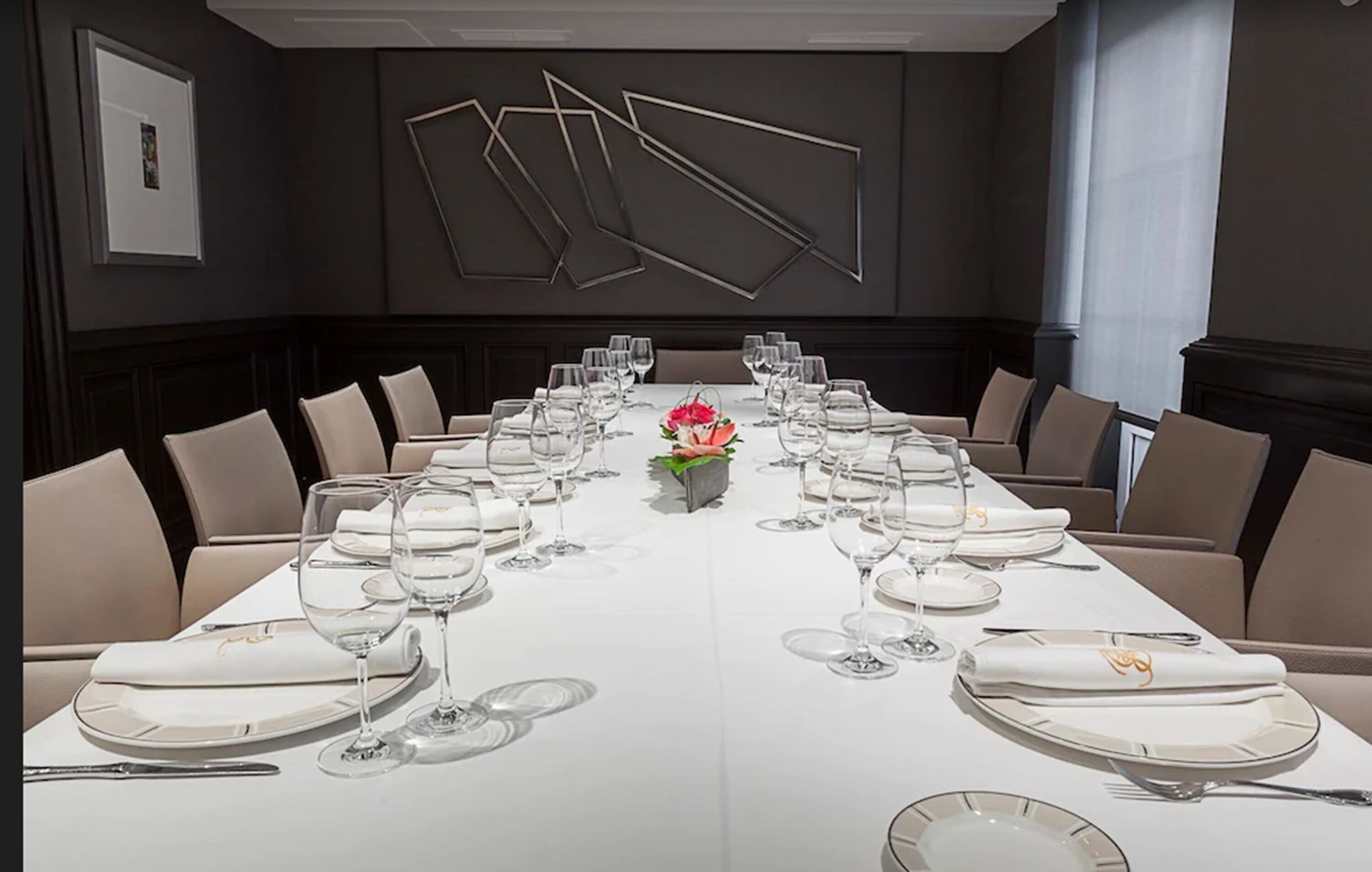 A long dining table set for a formal meal with multiple chairs, a centerpiece, and a modern wall design.