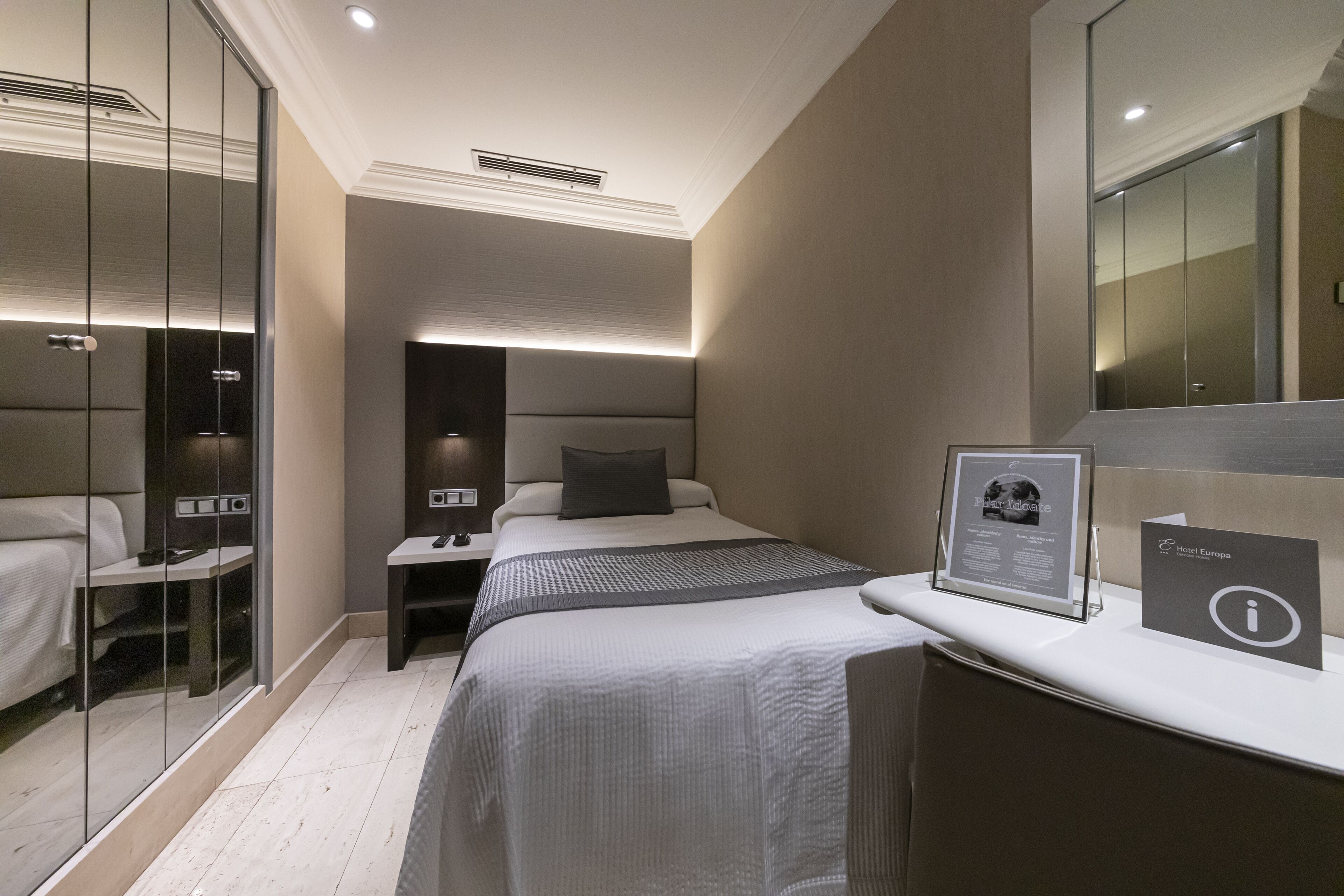 A modern hotel room with a large bed, bedside tables, a mirror, and a framed informational card.