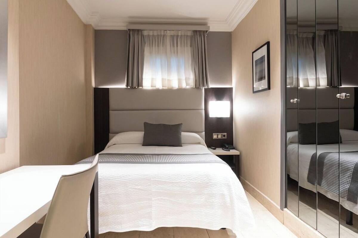 Quarto de hotel com cama, mesa de cabeceira, criadeira e armário com espelho.