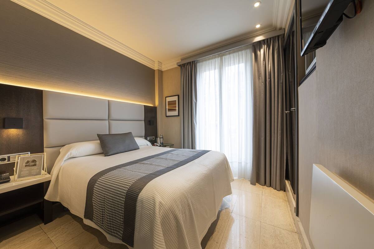Quarto de hotel moderno com uma cama grande, mesas de cabeceira, uma televisão de tela plana e um amplo janelão com cortinas.