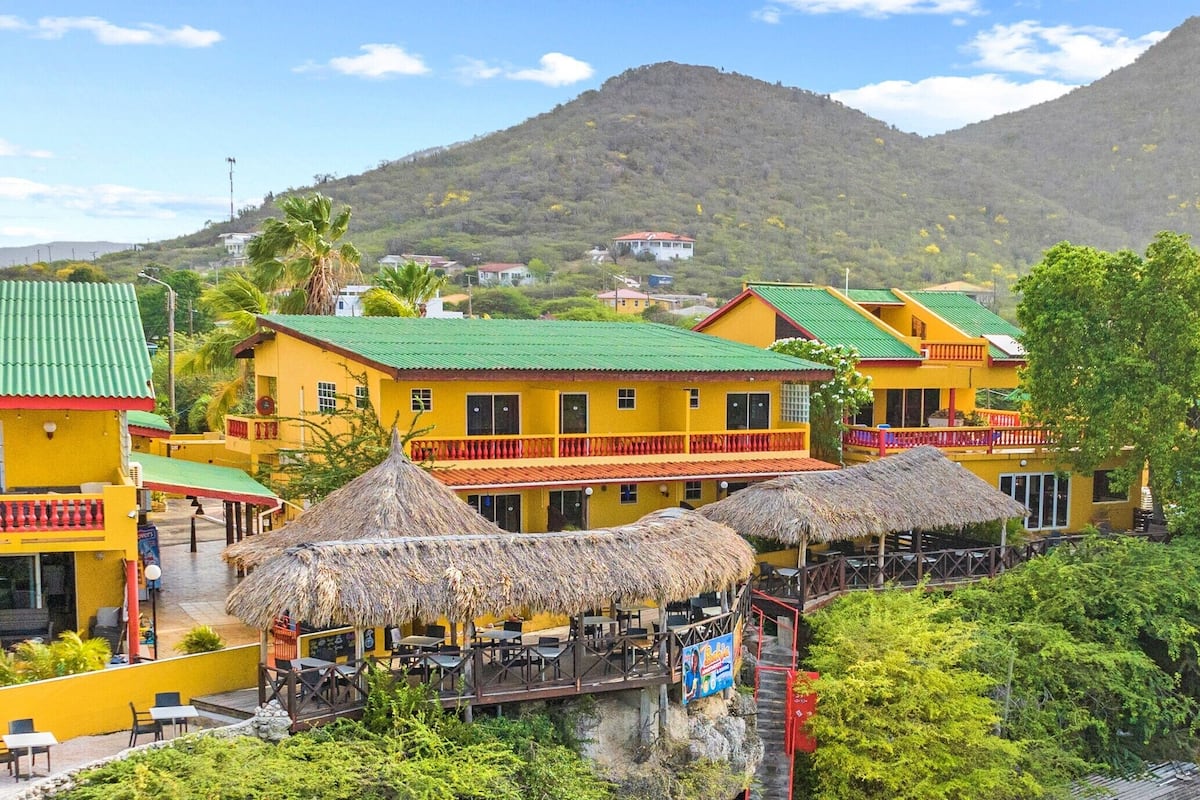 Un edificio amarillo con techo verde y techo de paja, con asientos exteriores y montañas al fondo.