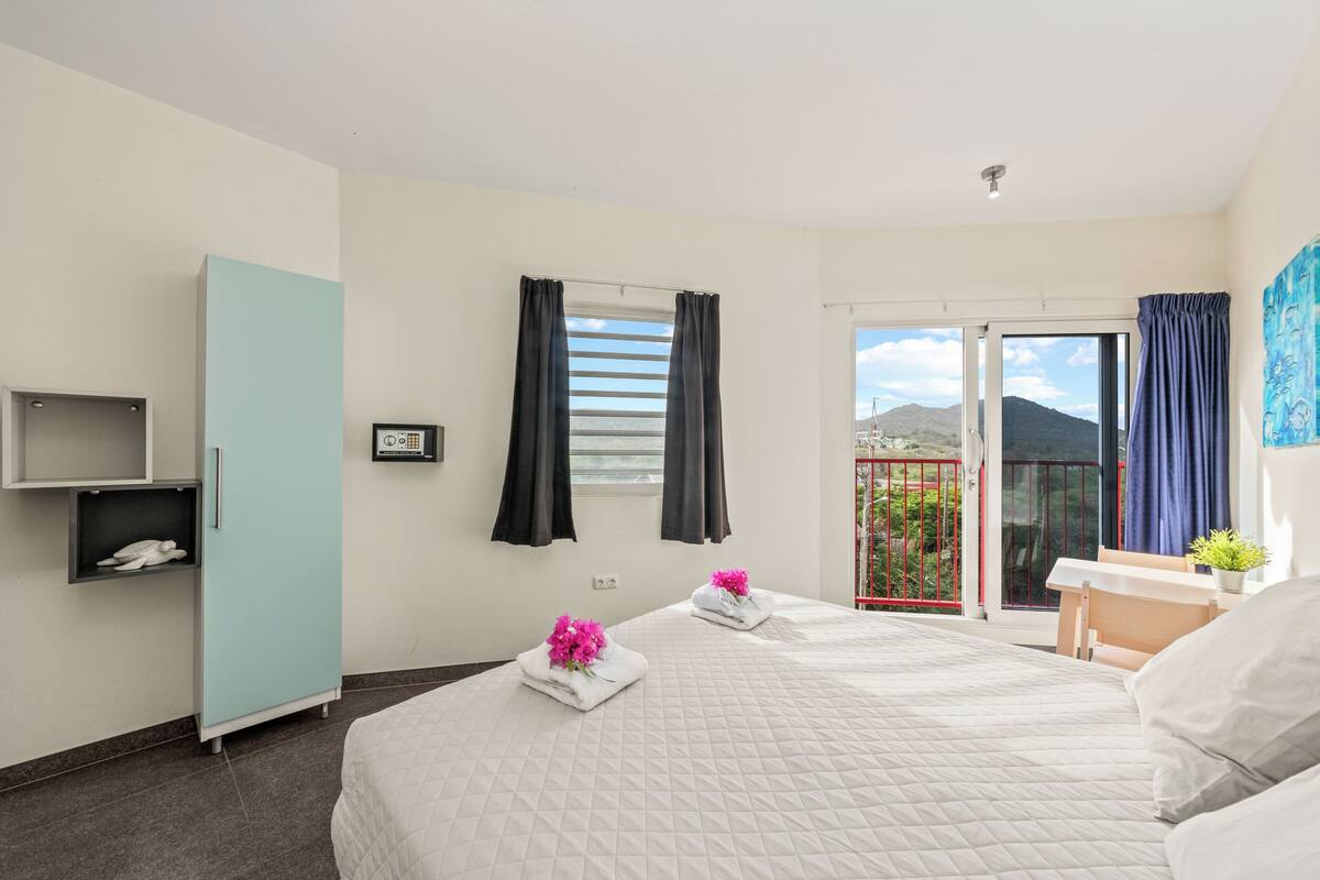 Habitación de hotel con cama, armario, estante pequeño, ventana con cortinas y balcón con vistas a las montañas.