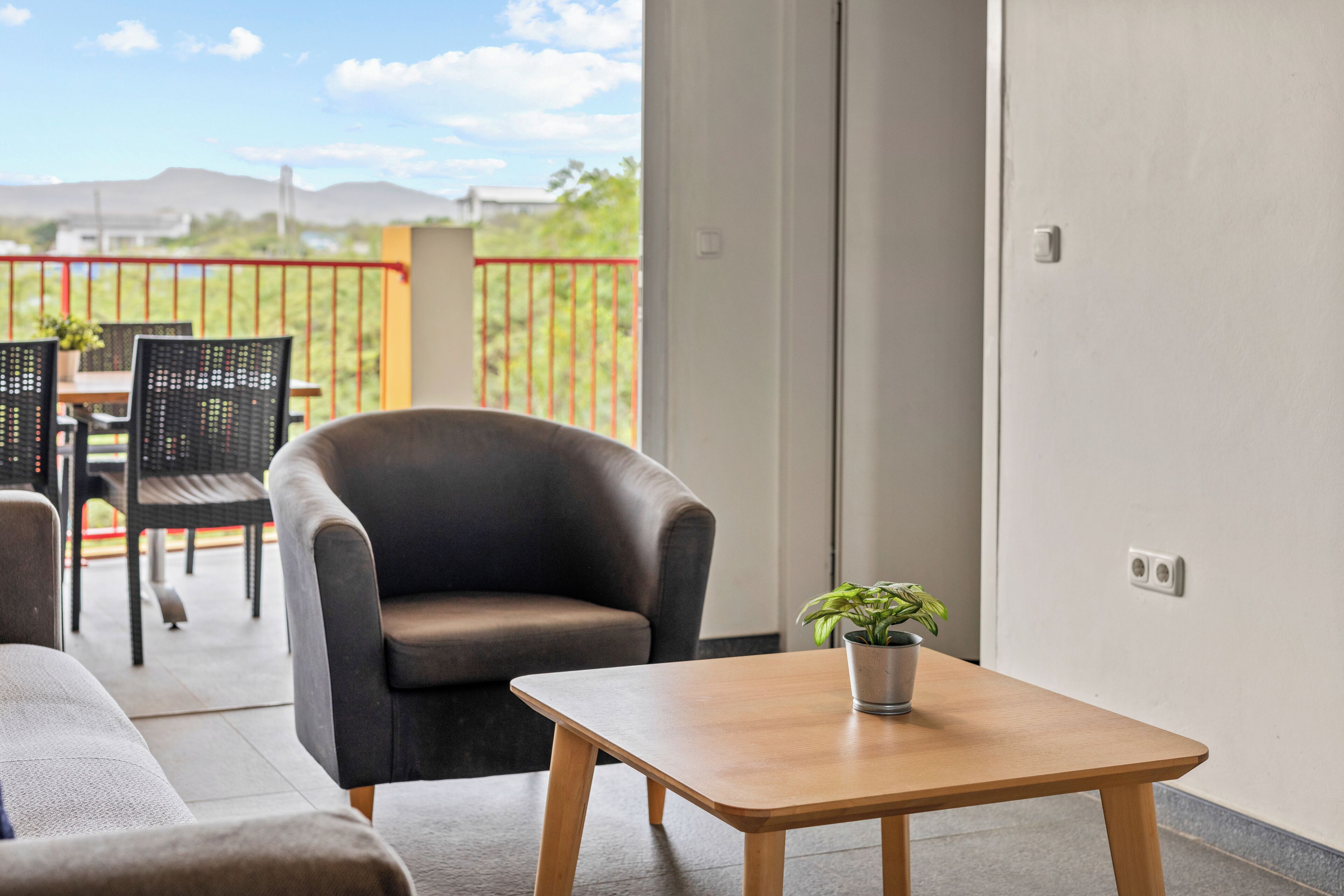 A modern living room with a sofa, armchair, and a small table with a plant. There is a balcony with outdoor furniture and a view of the mountains.