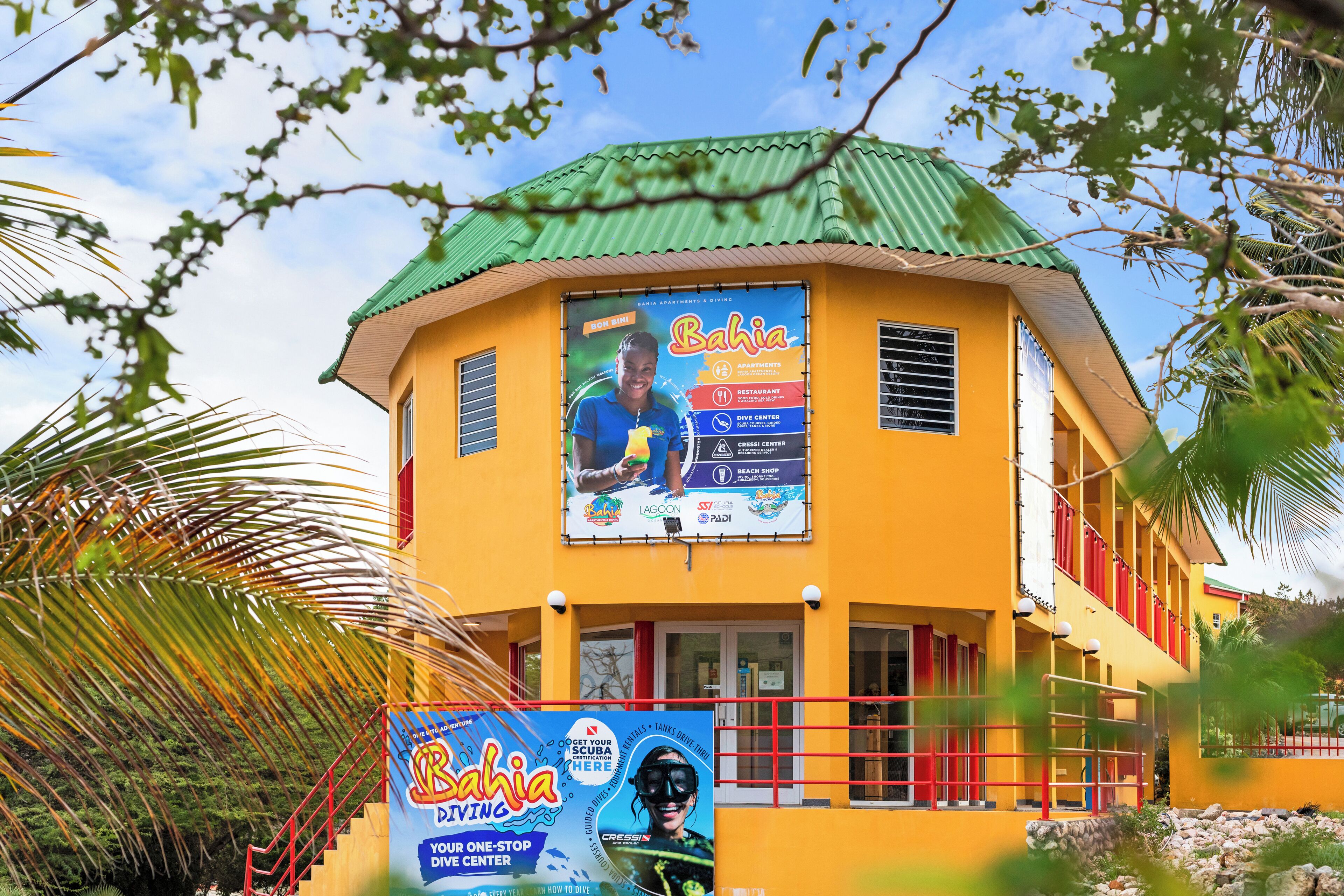 Un bâtiment jaune surmonté d’un toit vert, avec une grande enseigne publicitaire pour le centre de plongée Bahia Dive Center et, à proximité, le magasin de plongée de Rachael.