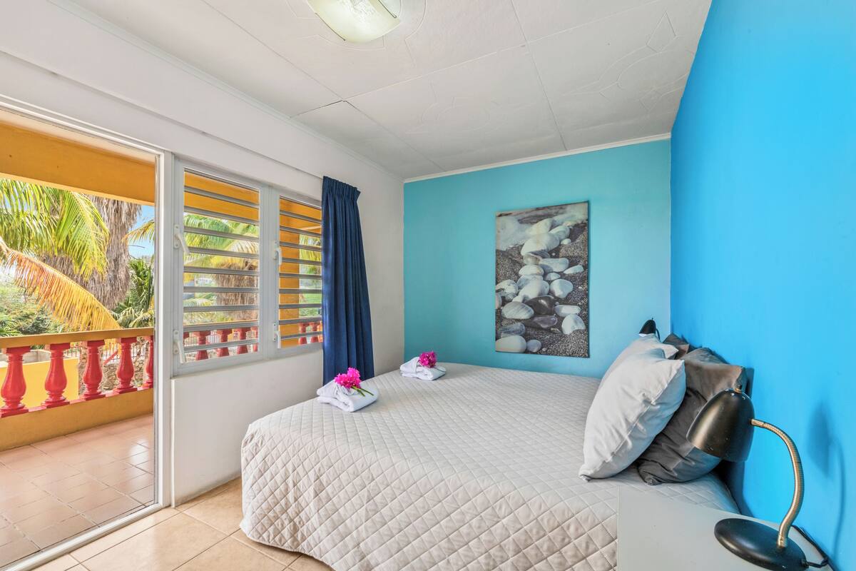 Un dormitorio con cama, un cuadro, una lámpara de escritorio y vistas a palmeras a través de una puerta de balcón.