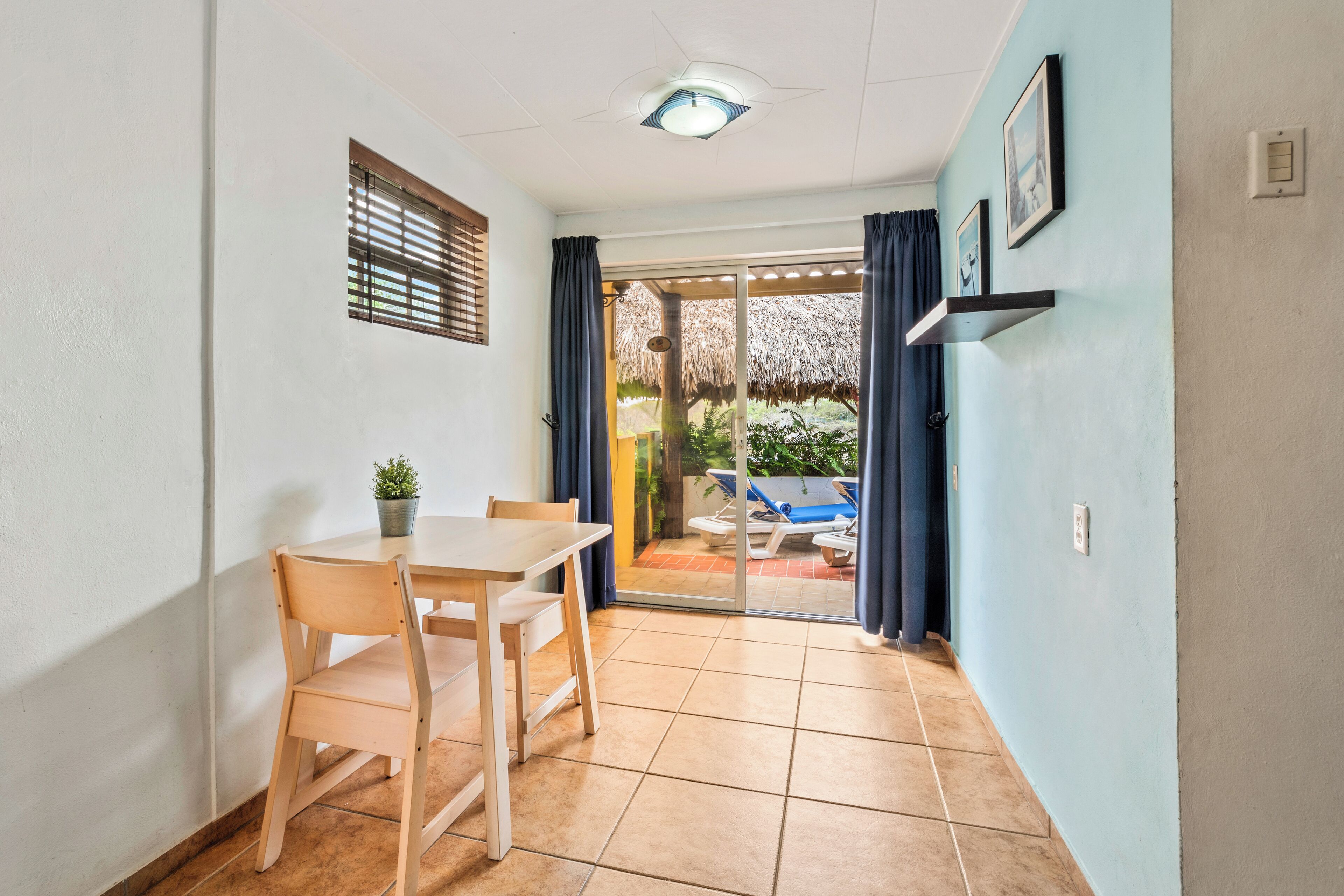 A room with a dining table and chairs, a small potted plant, a ceiling light, a sliding glass door leading to an outdoor area with a pool and umbrellas.