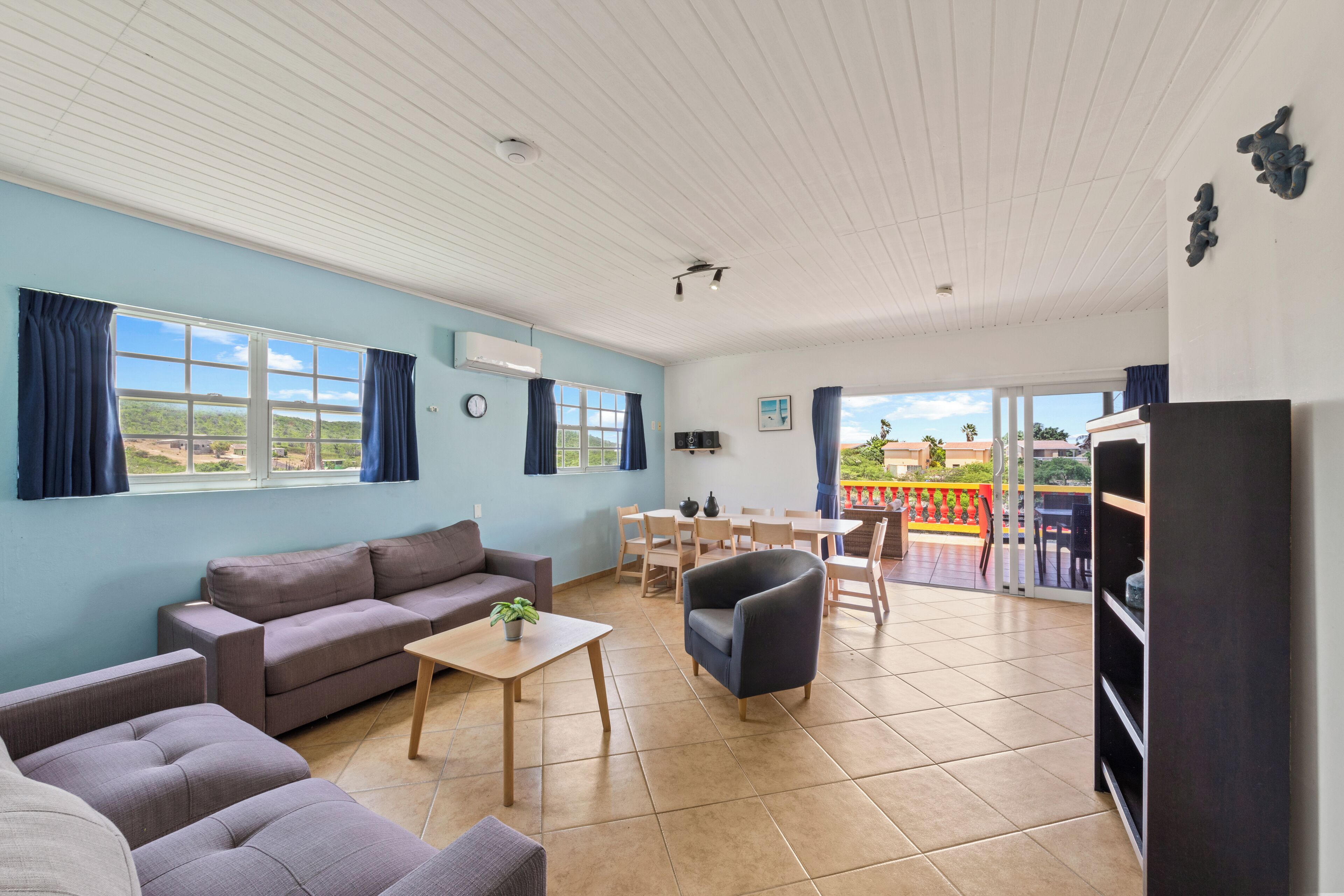 A spacious living area with a sofa, armchairs, a small table, and a balcony with a red railing.