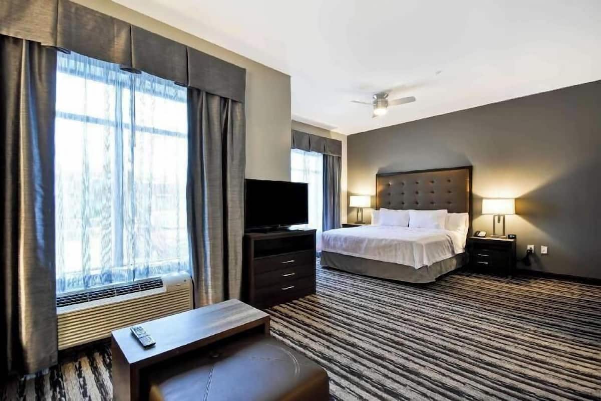 Habitación de hotel con cama, televisión, escritorio, silla, mesita de noche, lámpara y ventana con cortinas.