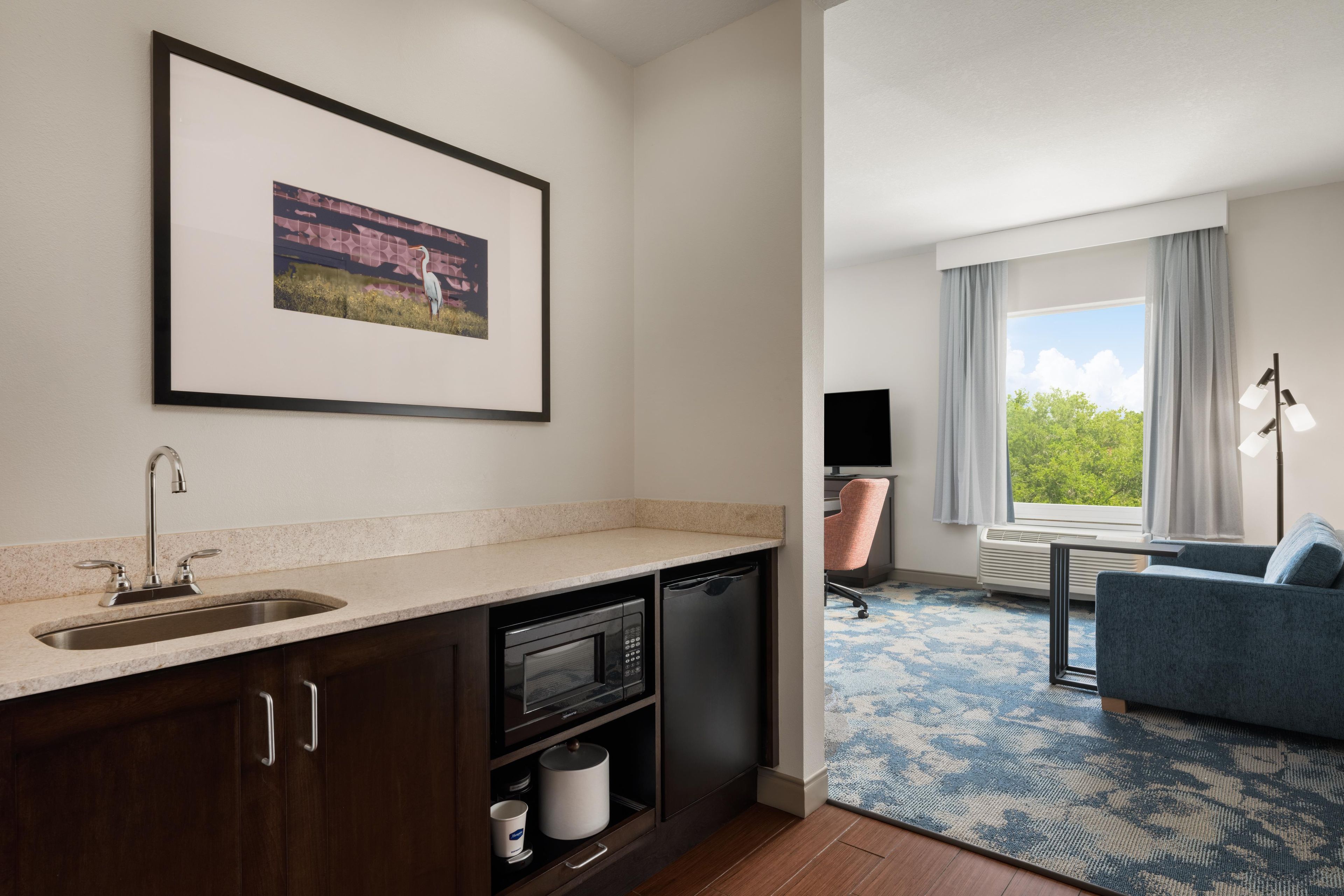Een moderne hotelkamer met kitchenette, een ingelijst kunstwerk aan de muur, een groot raam met gordijnen, een blauwe bank en een kamerbreed tapijt.