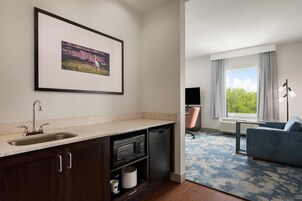 Una habitación de hotel moderna con kitchenette, un cuadro enmarcado en la pared, un ventanal con cortinas, un sofá azul y una alfombra estampada.