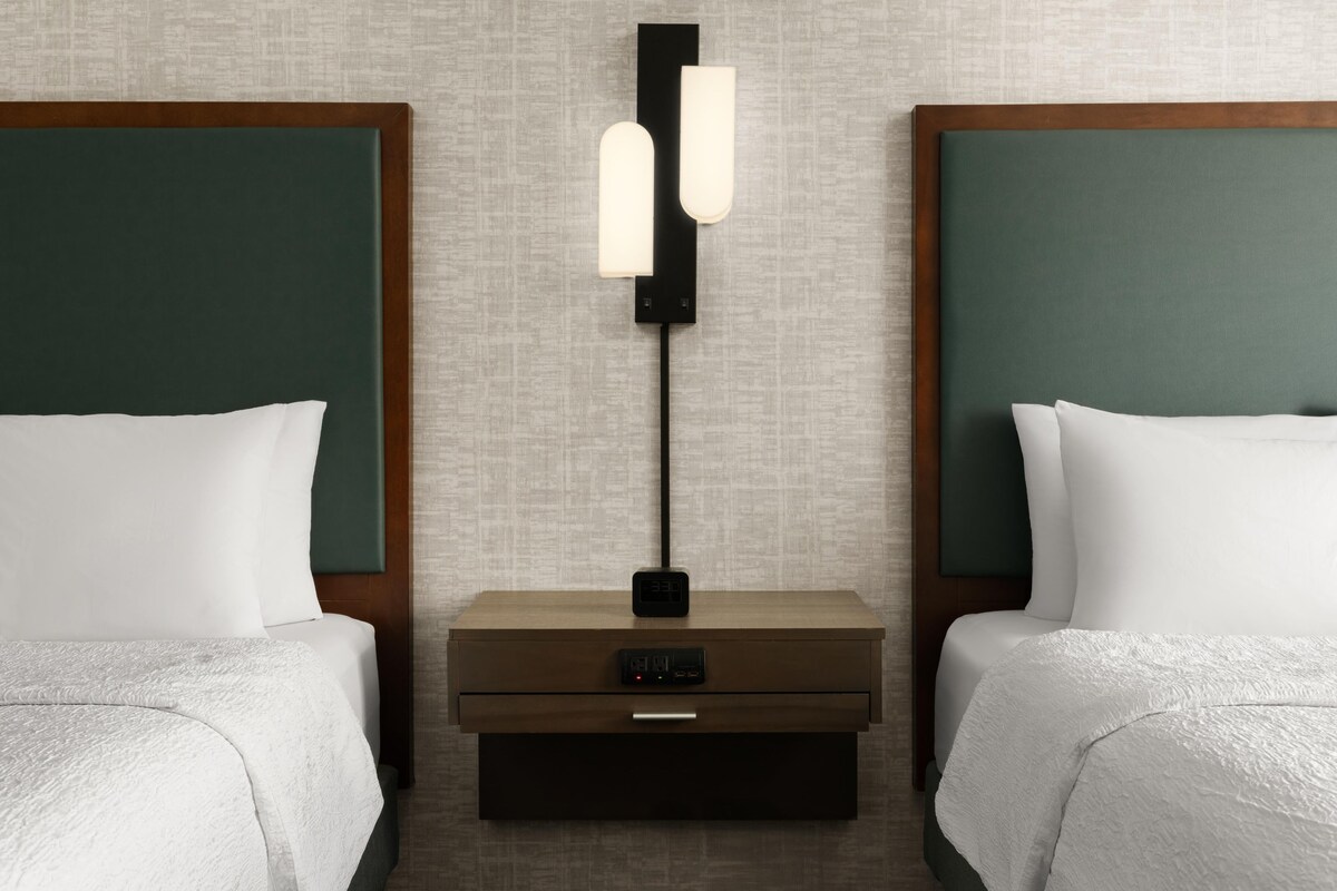 Een hotelkamer met twee bedden, een nachtkastje met een lamp en een digitale klok.