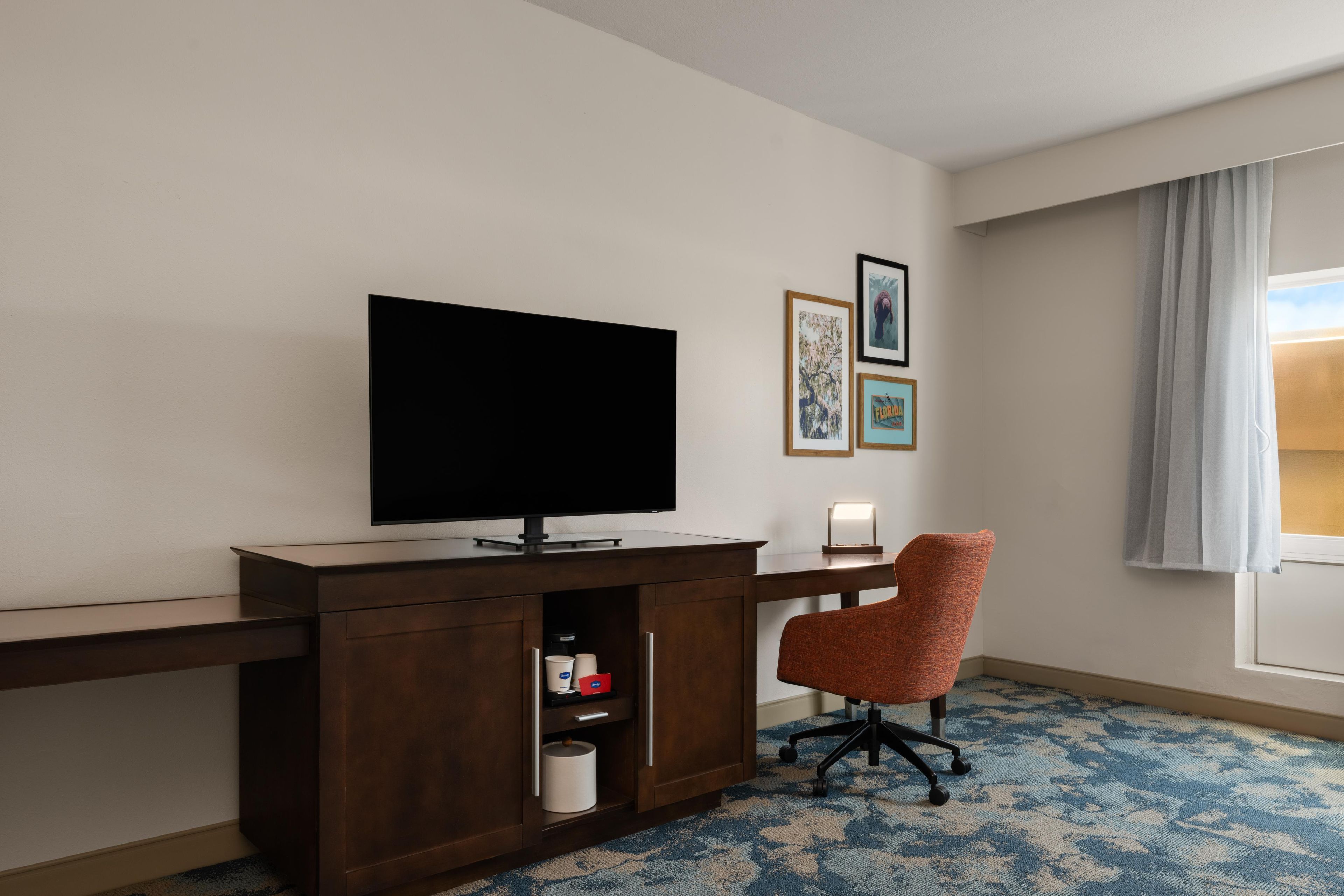 Quarto de hotel com televisão de tela plana, escrivaninha, cadeira, abajur e quadros na parede.
