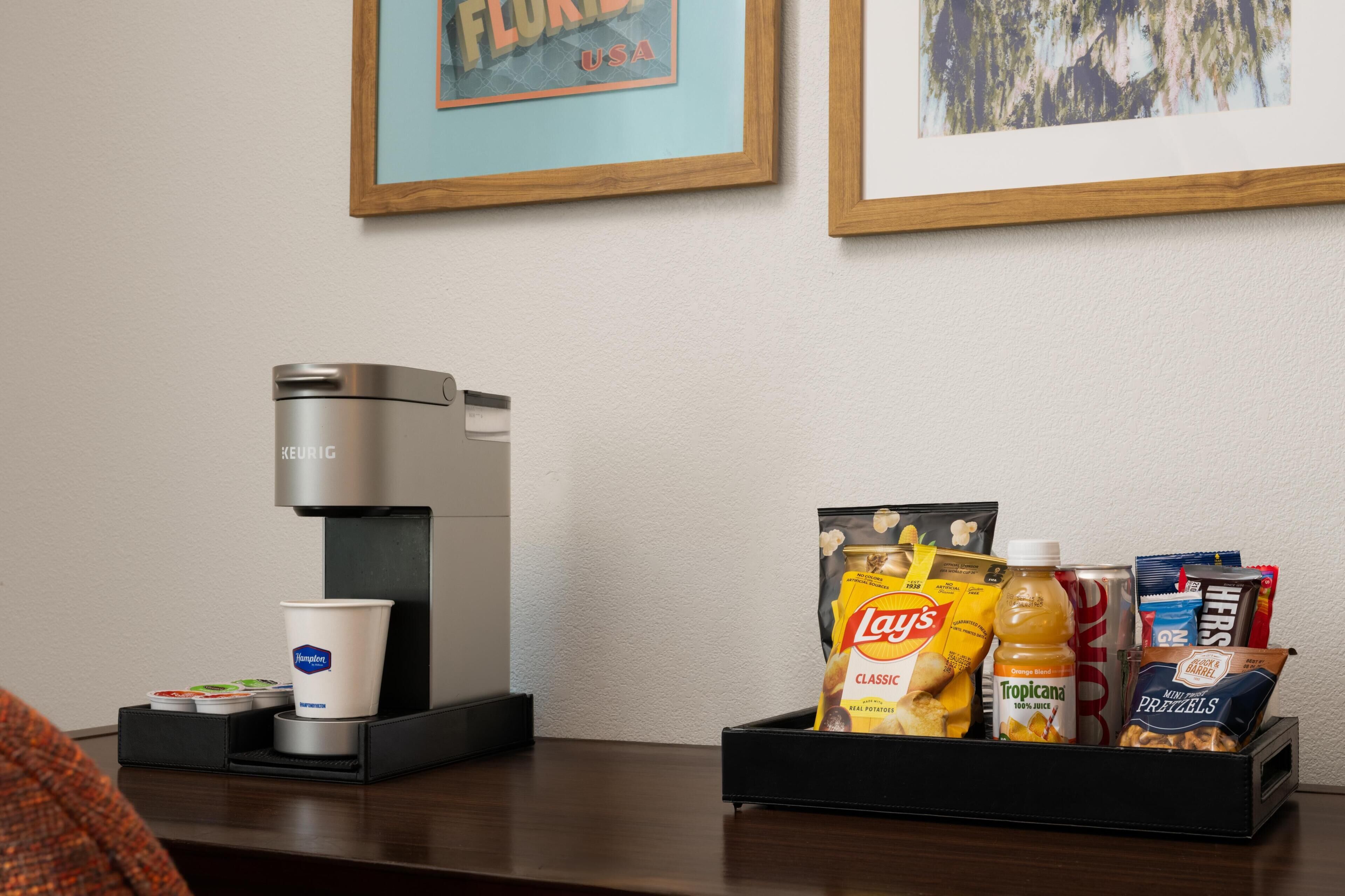 A coffee machine, a cup, and a snack tray with chips, juice, and pretzels on a table.