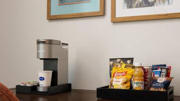 A coffee machine, a cup, and a snack tray with chips, juice, and pretzels on a table.