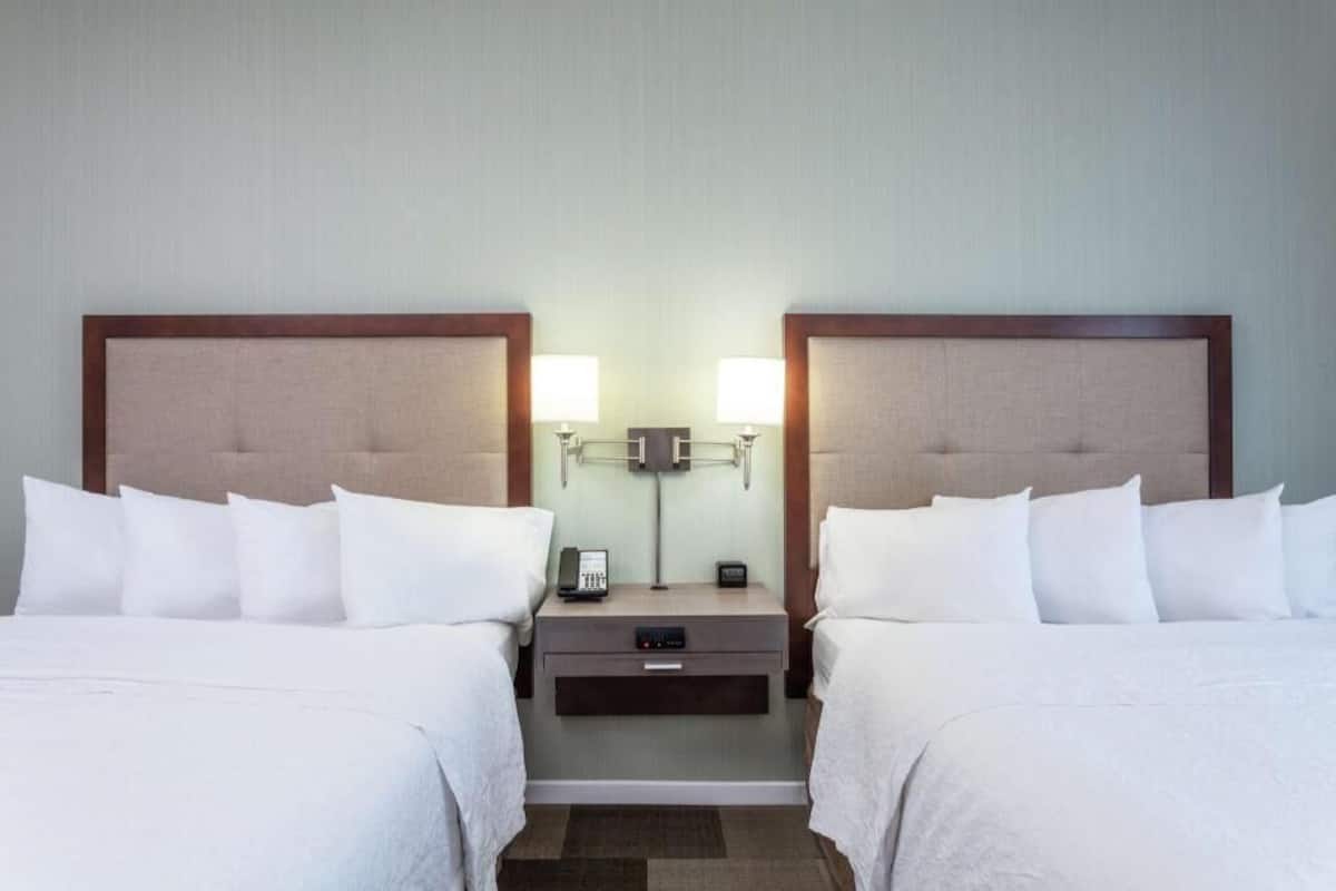 A hotel room with two beds, a nightstand with a lamp, and a small table with a phone and a remote control.