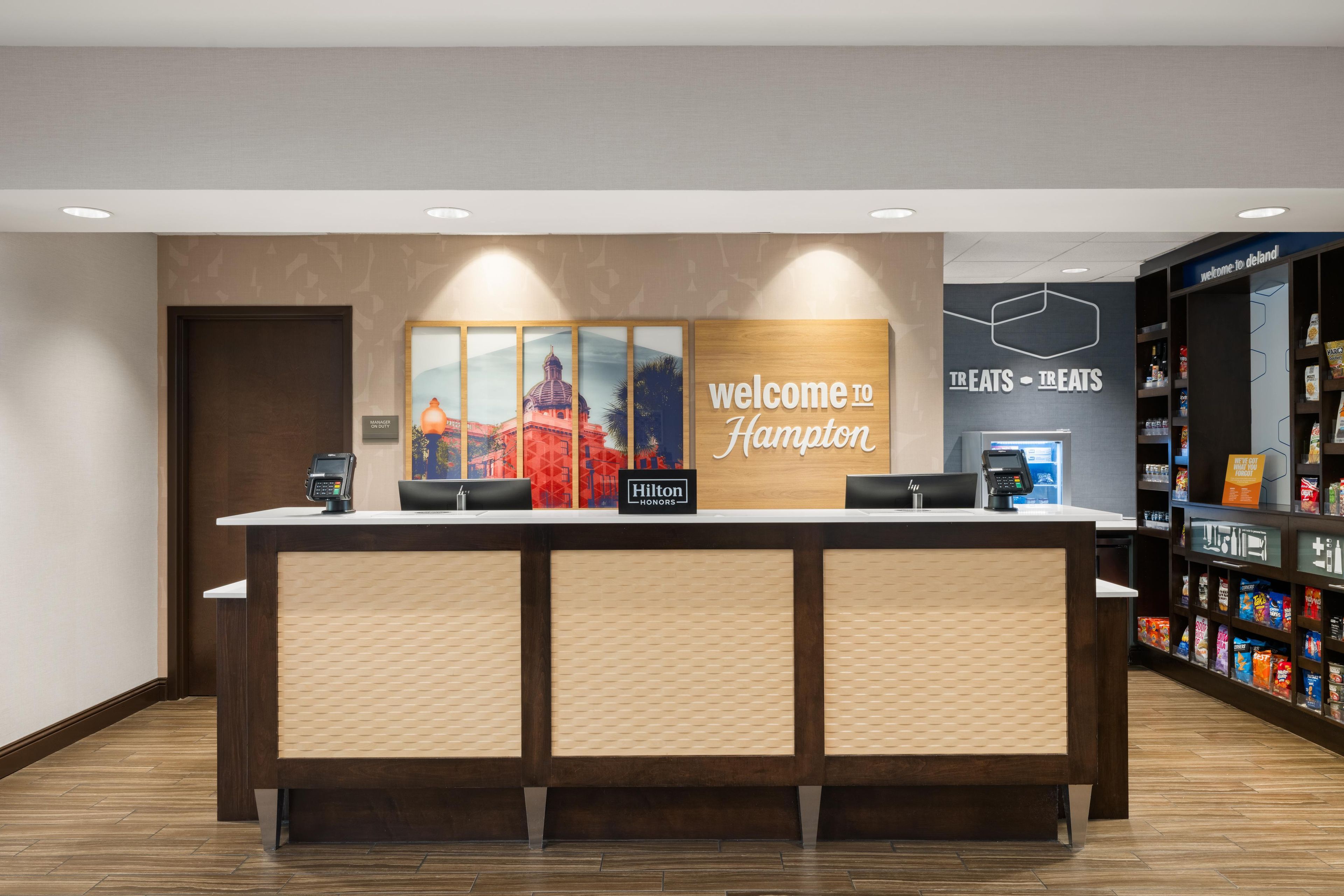 A hotel reception desk with a 'Welcome to Hampton' sign, a 'Manager on Duty' sign, and a 'Hilton Honors' sign.
