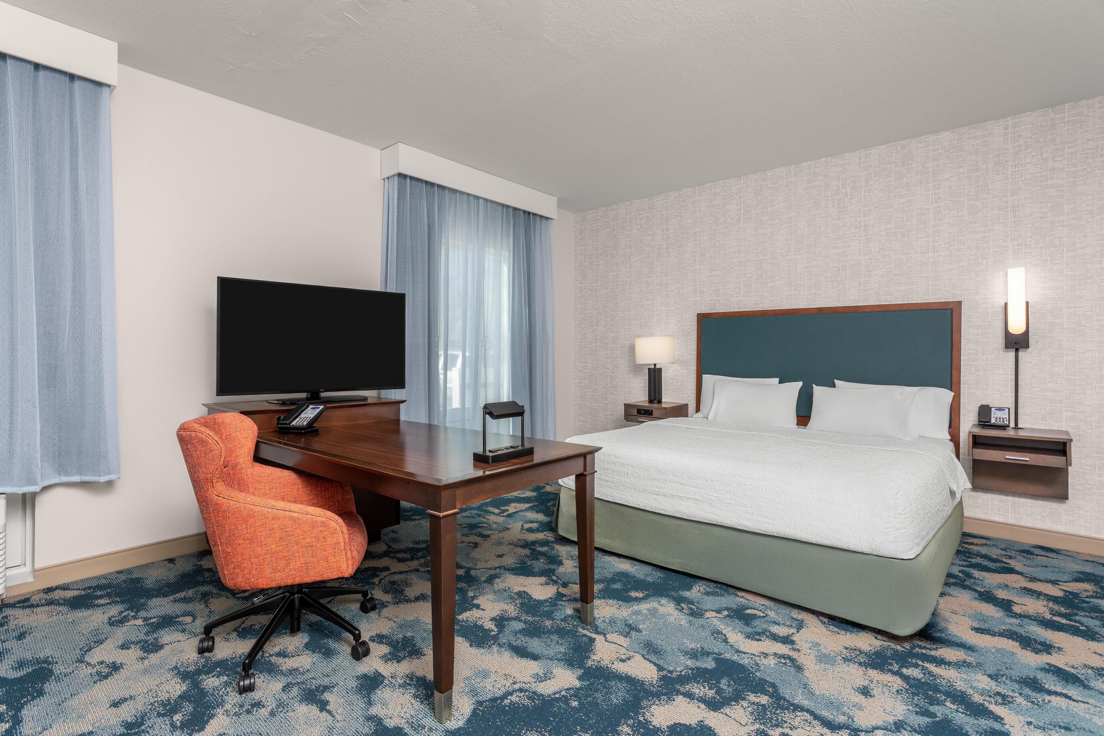 Et hotelværelse med seng, skrivebord med tv, stol, lampe og et natbord.
