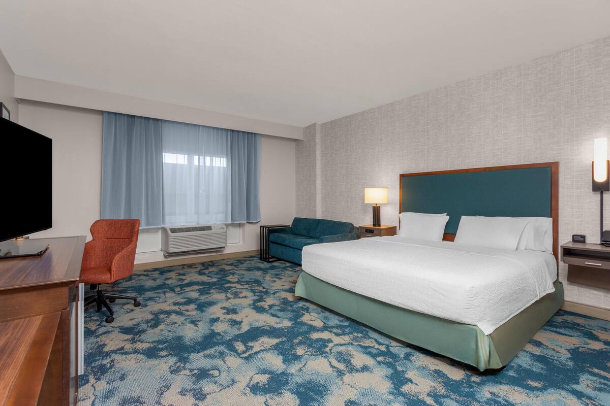A hotel room with a bed, a desk, a TV, a chair, a sofa, and a window with curtains.