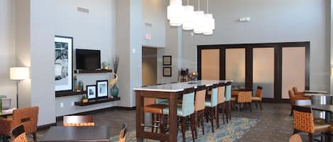 A hotel lobby with a long communal table, several chairs, a television, framed pictures, and a patterned carpet.