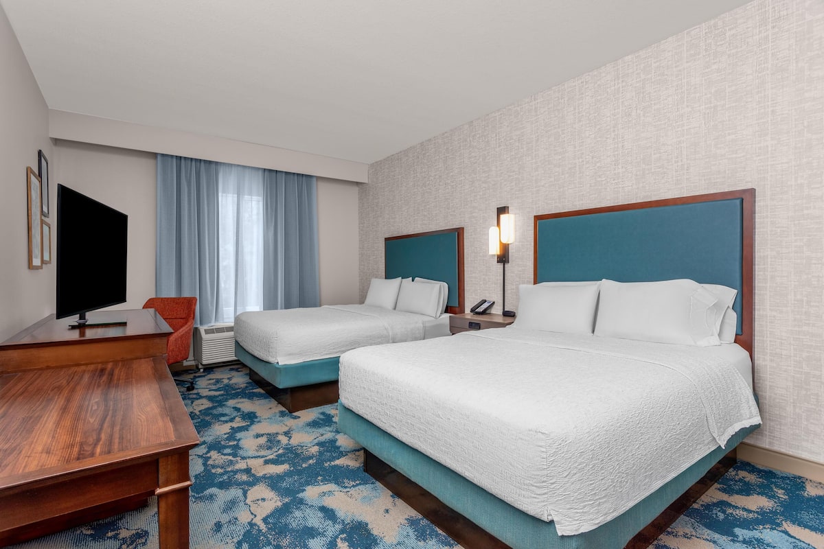 A hotel room with two beds, a desk, a TV, and a window with curtains.