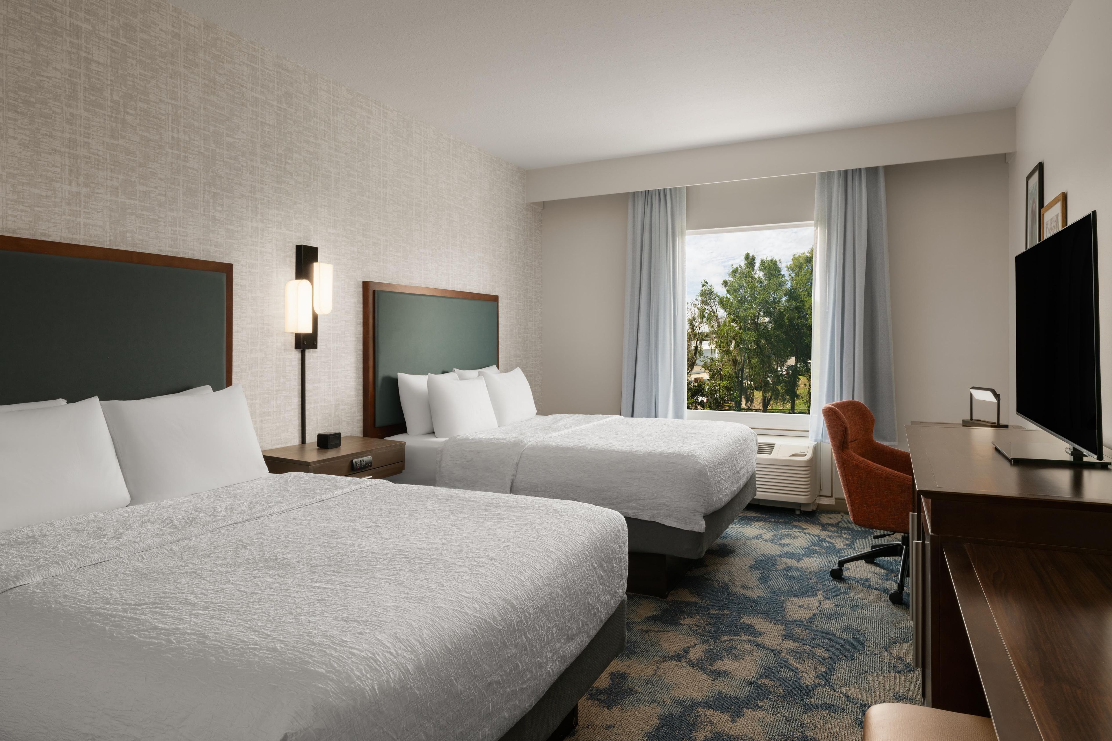A hotel room with two beds, a desk, a TV, and a window with a view of trees.