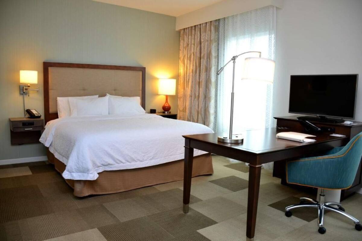 A hotel room with a bed, bedside lamps, a desk with a chair, a television, and a window with curtains.