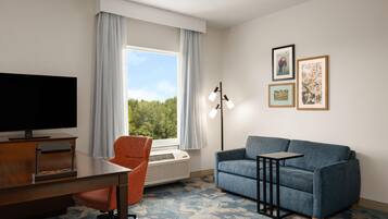 A hotel room with a television, a desk, a blue sofa, a chair, a standing lamp, and a large window with curtains.