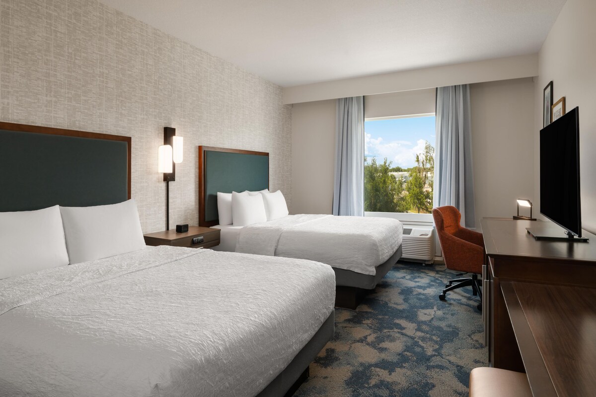 Een hotelkamer met twee bedden, een bureau, een stoel, een televisie en een raam met uitzicht op bomen.