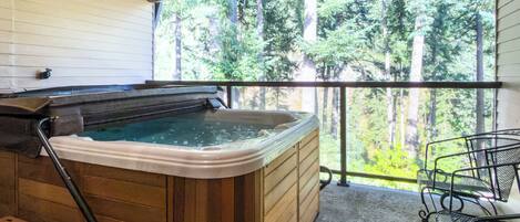 Een hottub met een houten buitenkant, een metalen stoel en uitzicht op een bos door een balkonhek.