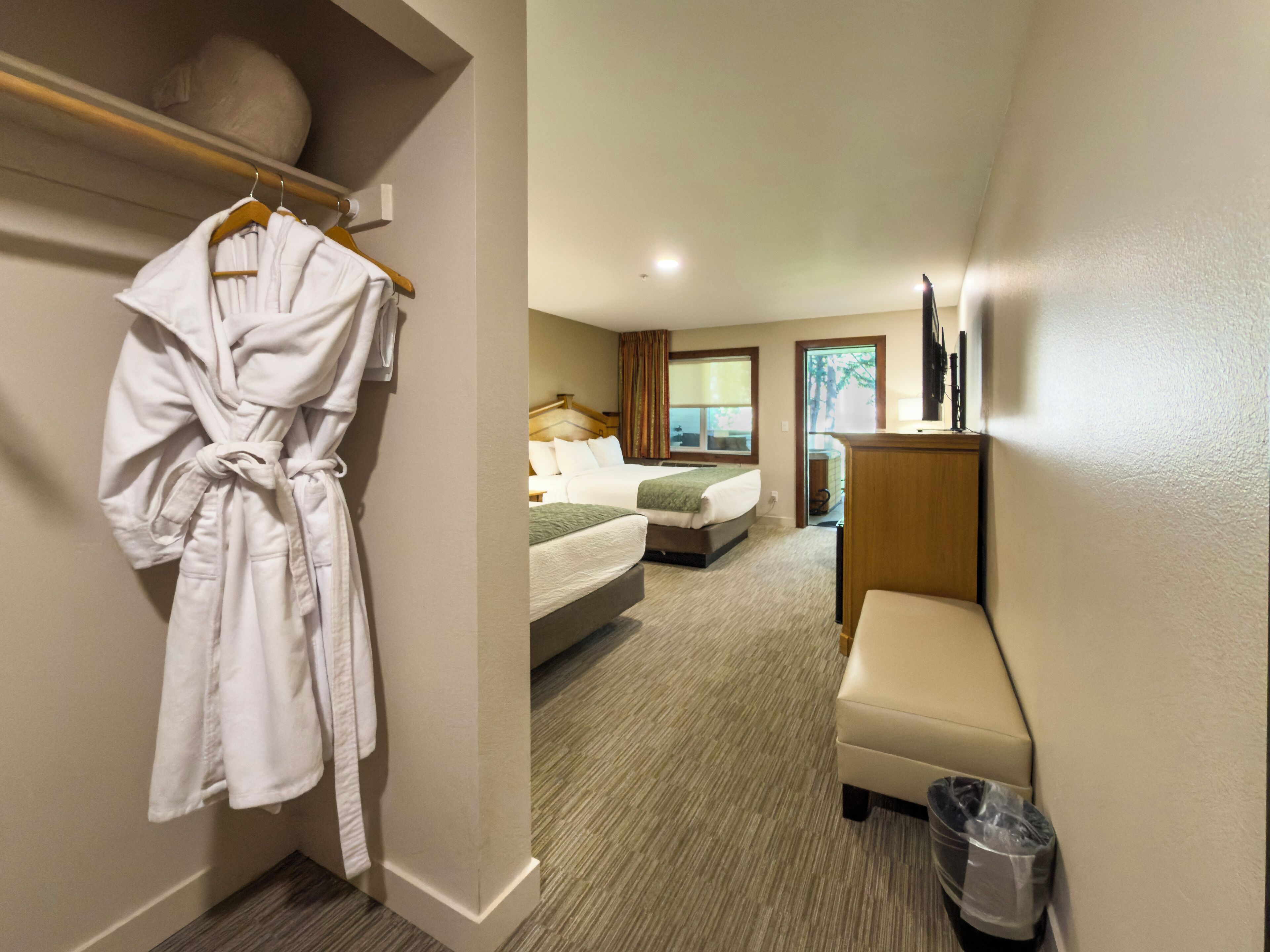 A hotel room with a bed, a bench, a robe hanging on a rack, and a trash bin.