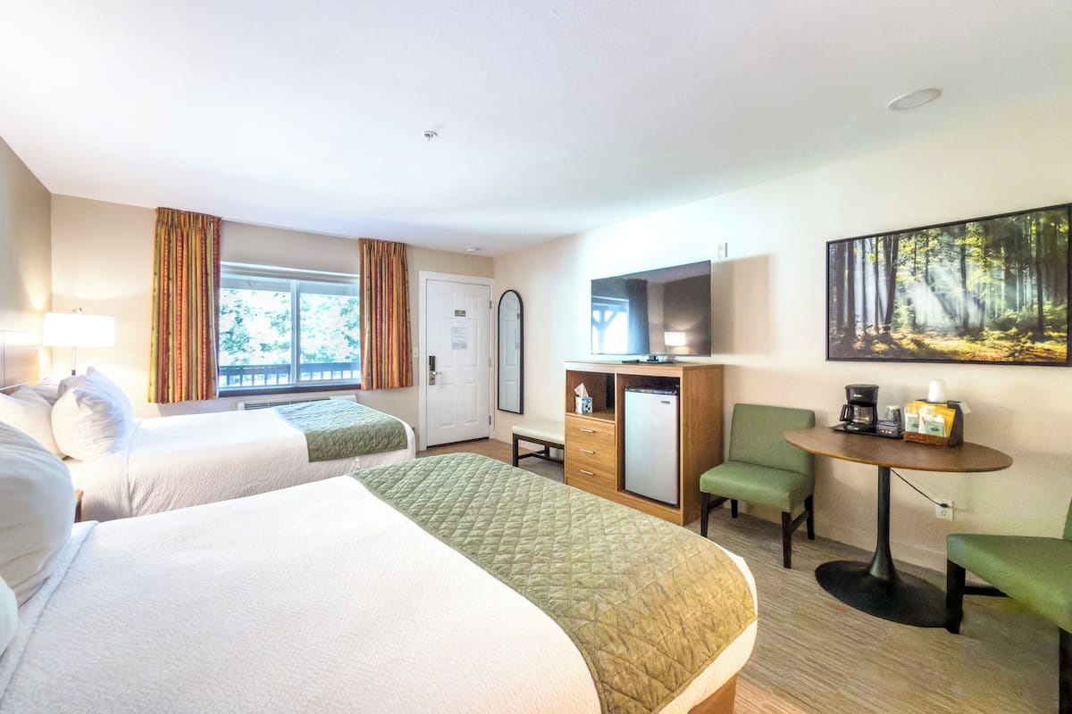 A hotel room with two beds, a desk with a computer, a TV, and a painting of a forest.