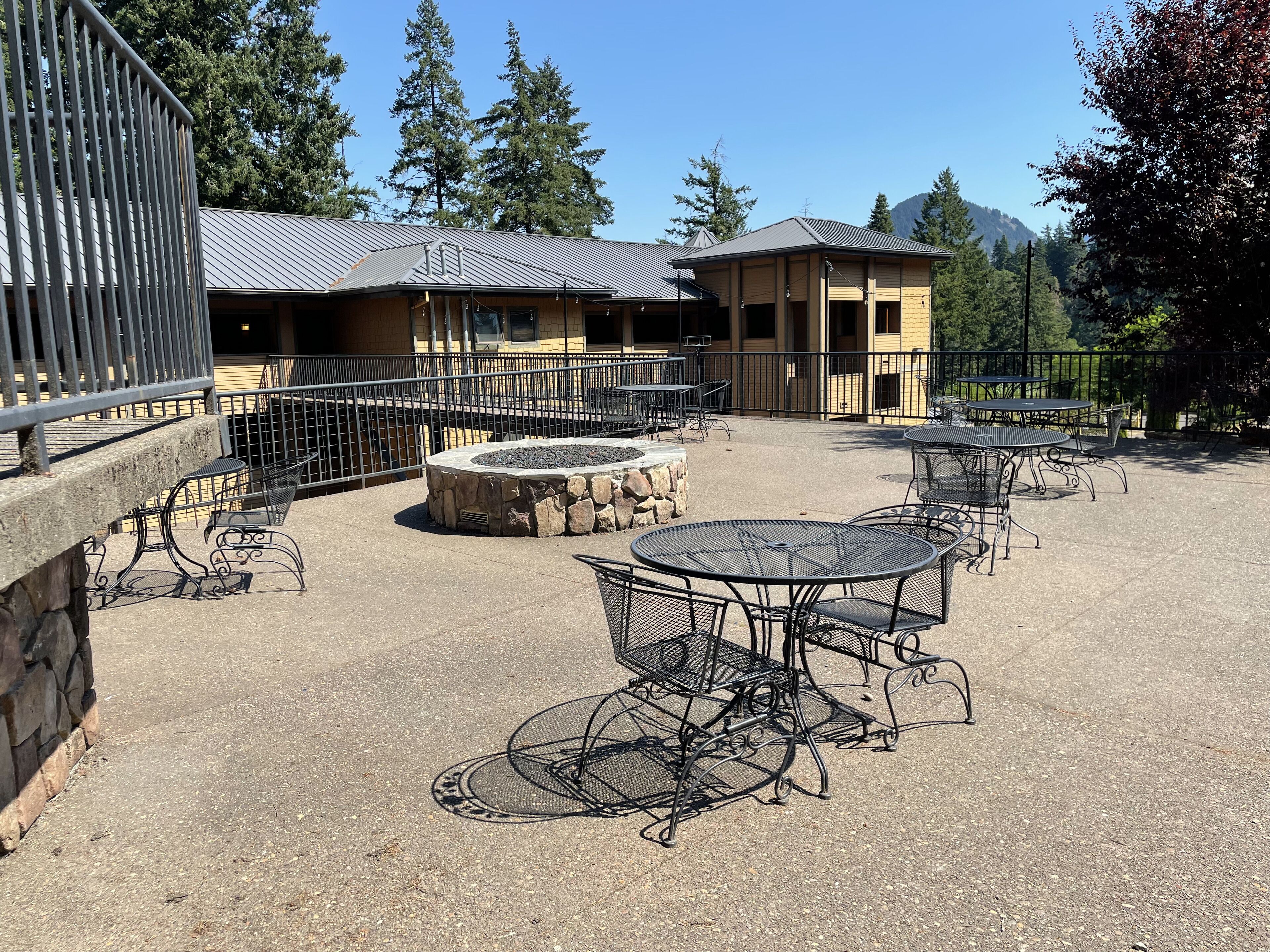Un patio al aire libre con mesas y bancos de metal, un hogar de piedra y un edificio con techo de metal al fondo.