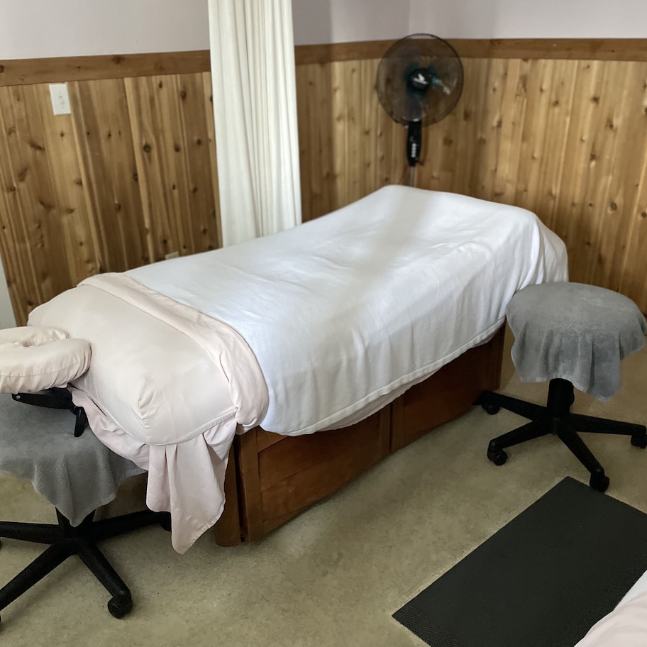 A treatment room with a wooden panel wall, a massage table, a footrest, a fan, and a small table with candles.