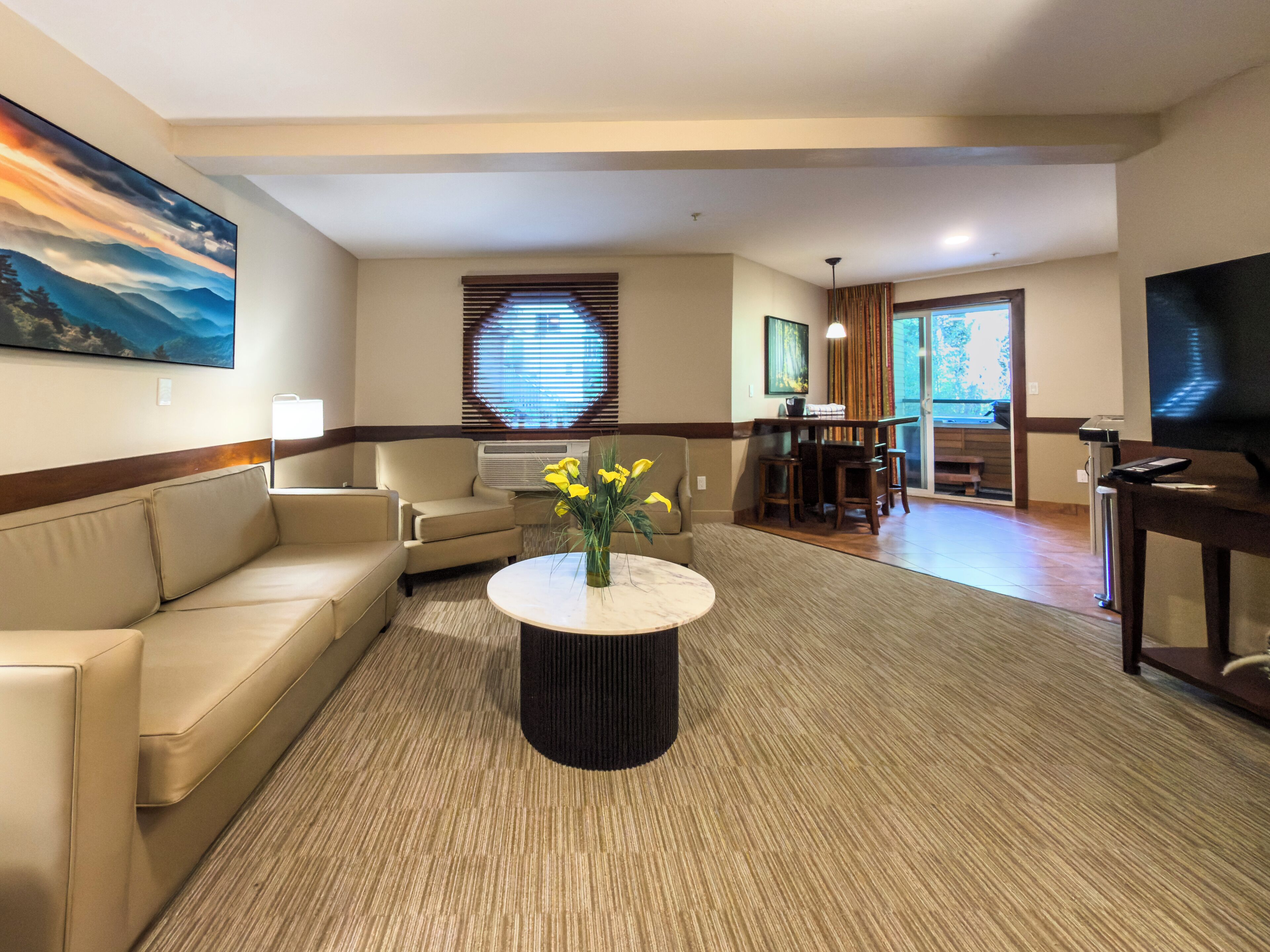 A hotel living room with a sofa, a coffee table with flowers, a TV, and a dining area with a table and chairs.