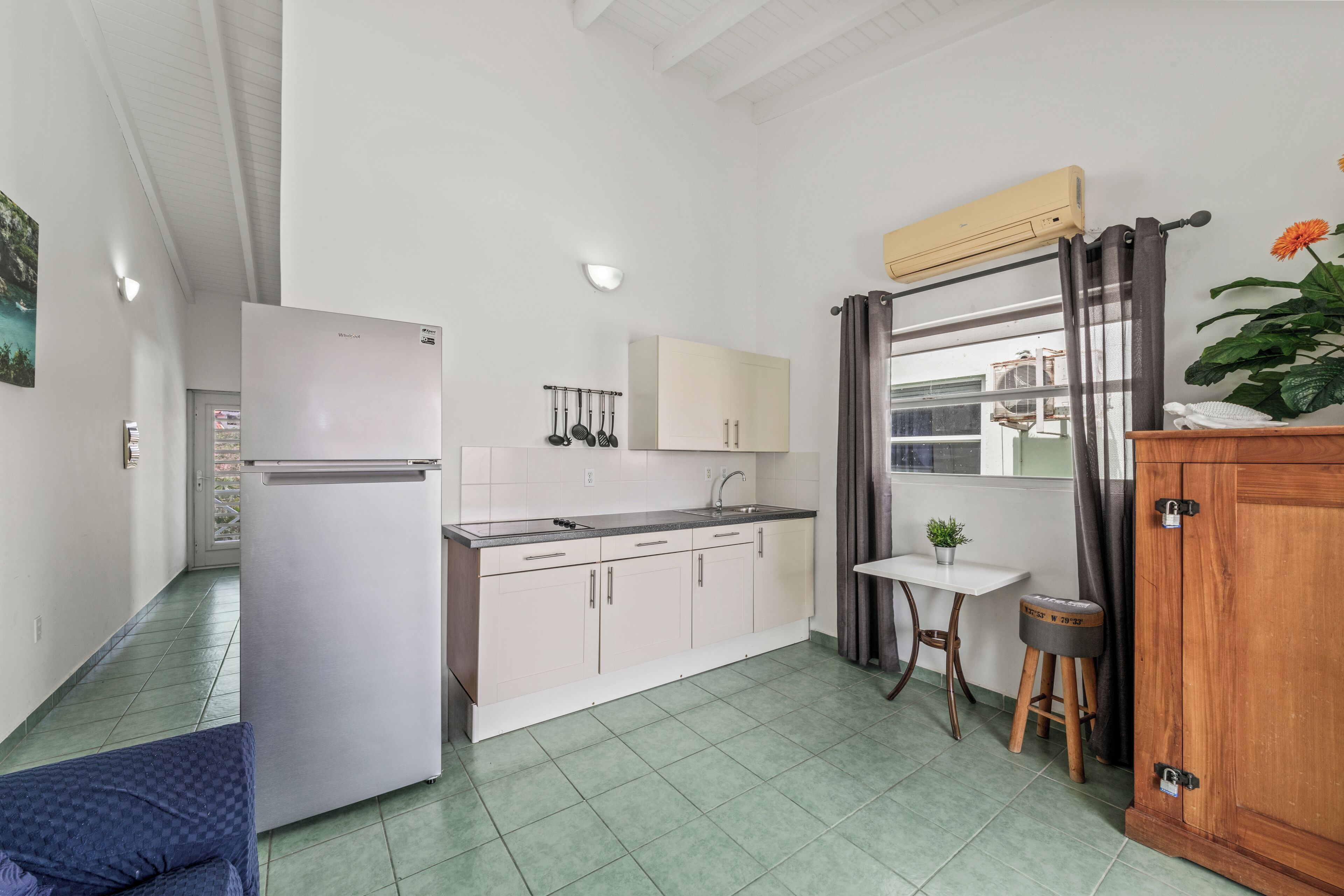 A compact kitchen with a refrigerator, sink, and cabinets. There is a small table with a potted plant and a stool.