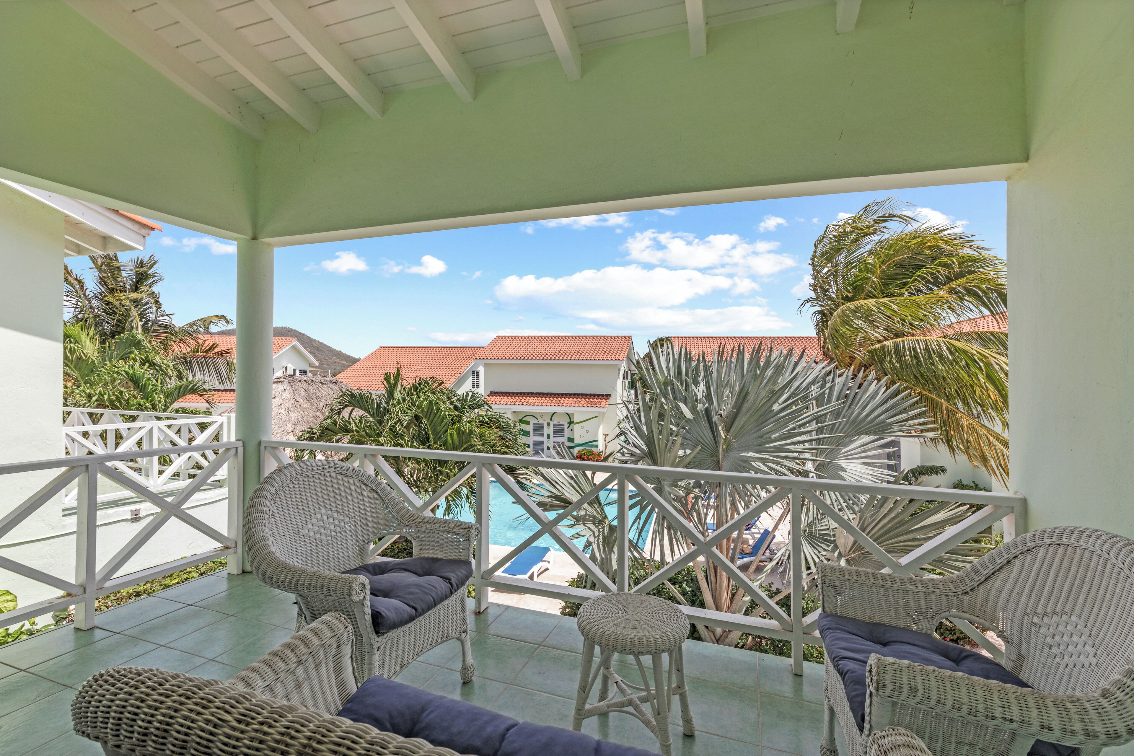 A covered balcony with wicker furniture, a pool, and a view of buildings and palm trees.
