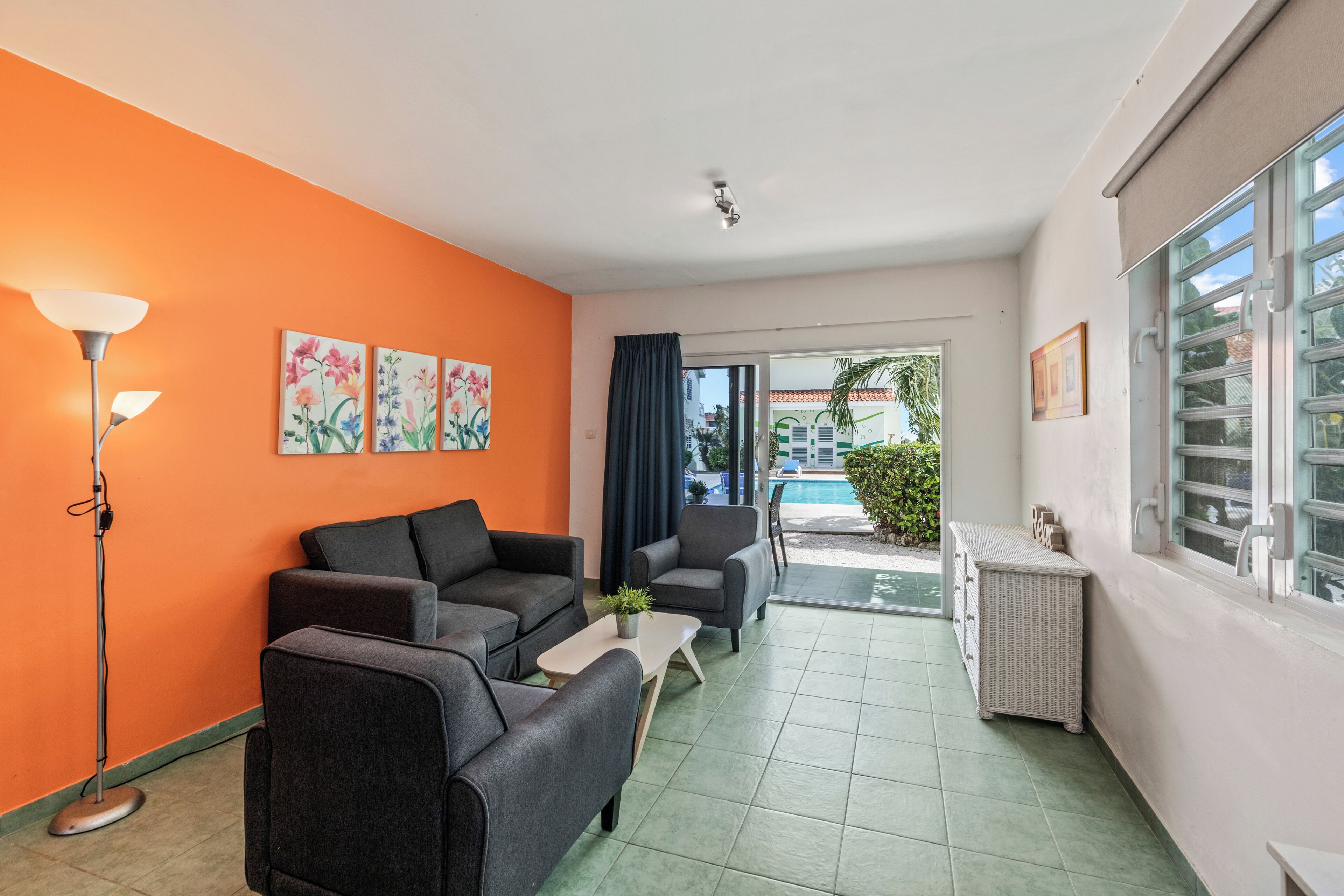Una habitación con sofá, sillones, una mesita, una lámpara de pie y un ventanal con vistas a una piscina y vegetación.