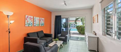 Una habitación con sofá, sillones, una mesita, una lámpara de pie y un ventanal con vistas a una piscina y vegetación.