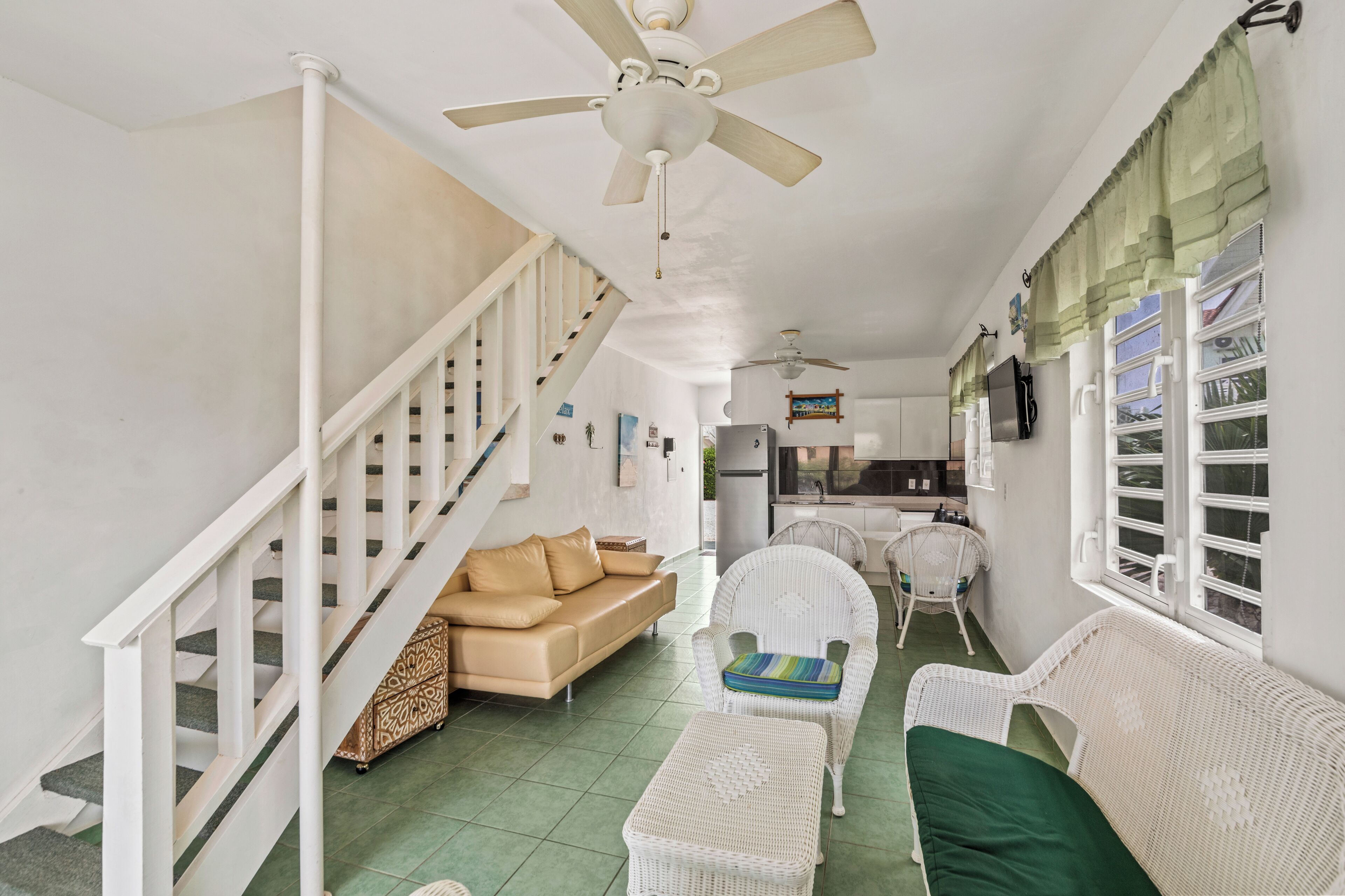 A spacious living area with a staircase, white wicker furniture, and a kitchen in the background.