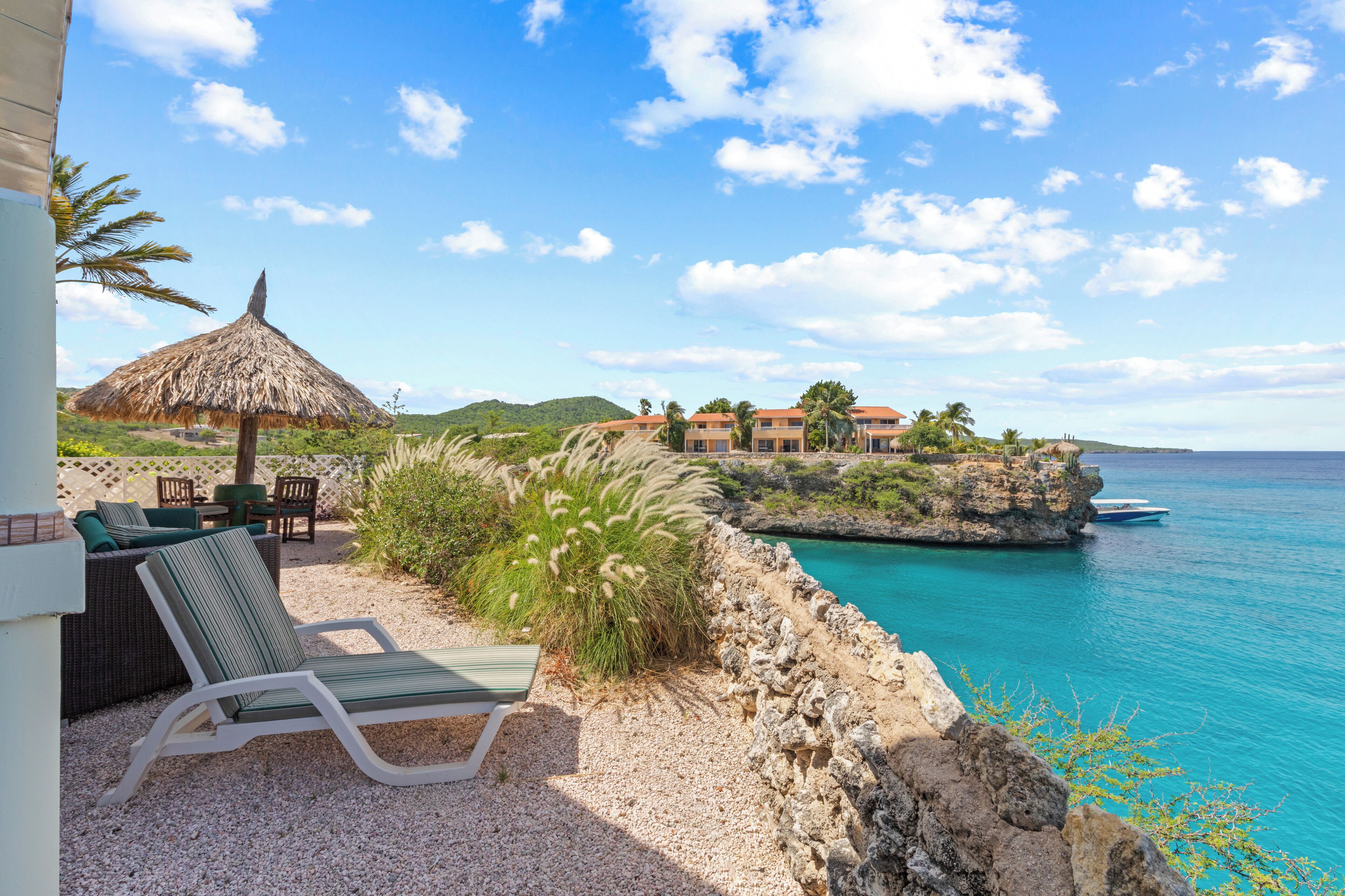 A coastal view with a thatched umbrella, outdoor seating, and a clear blue sea.